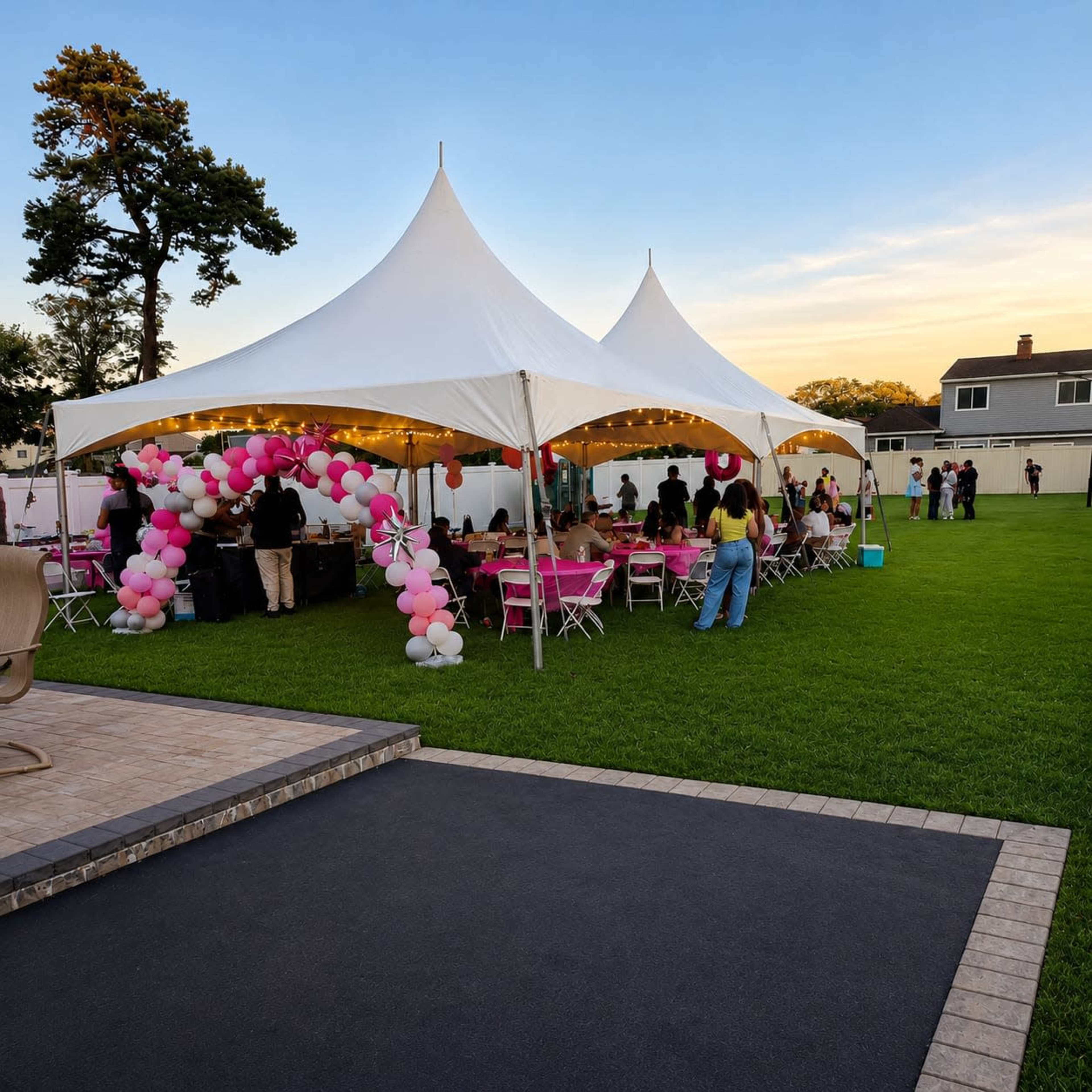 A large white tent filled with tables and chairs is set up on a grassy lawn, decorated with pink balloons, while guests socialize under a sunset sky.