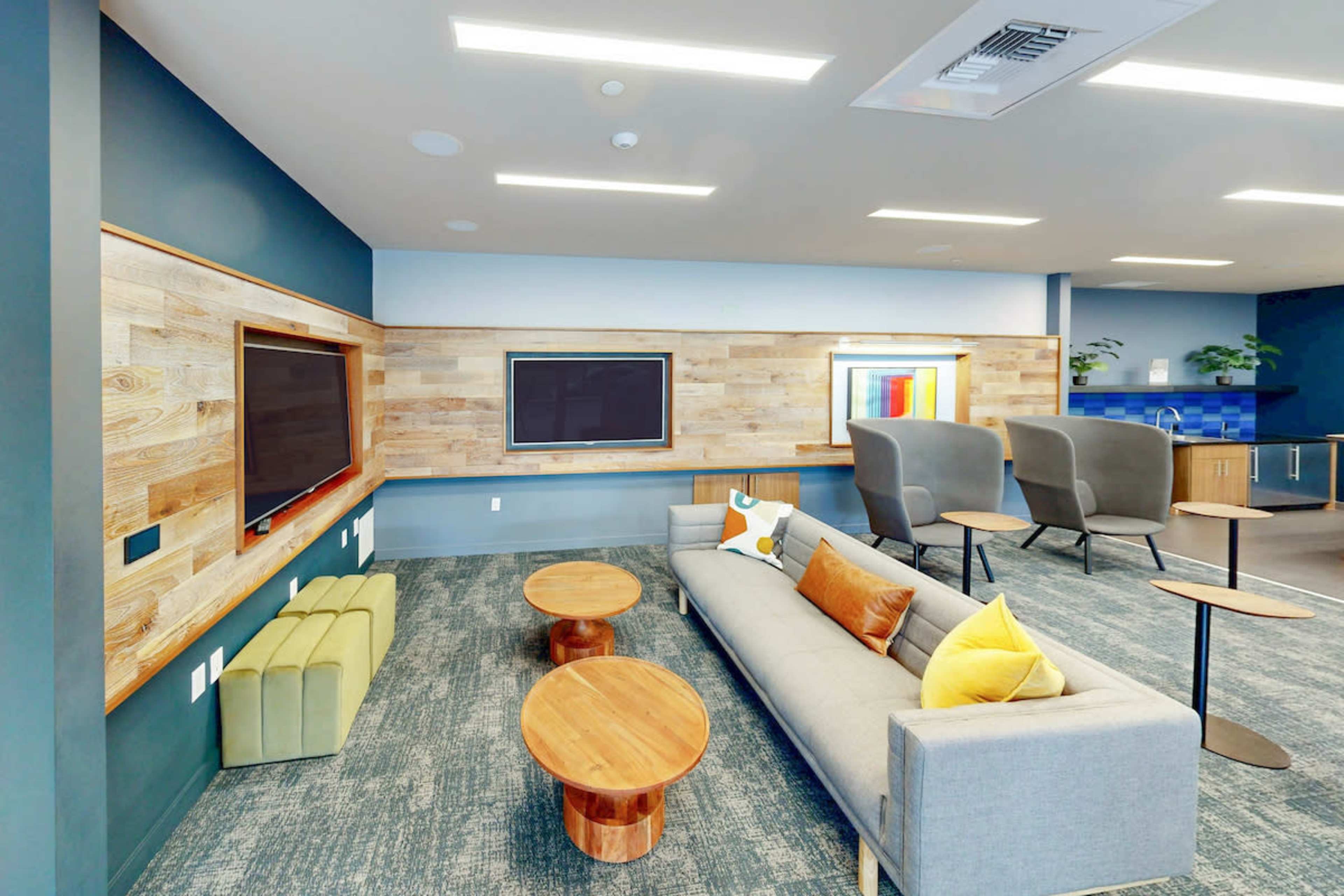 The image shows a modern lounge area featuring a gray sofa, wooden coffee tables, two large screens mounted on a wooden wall, and a kitchenette in the background.