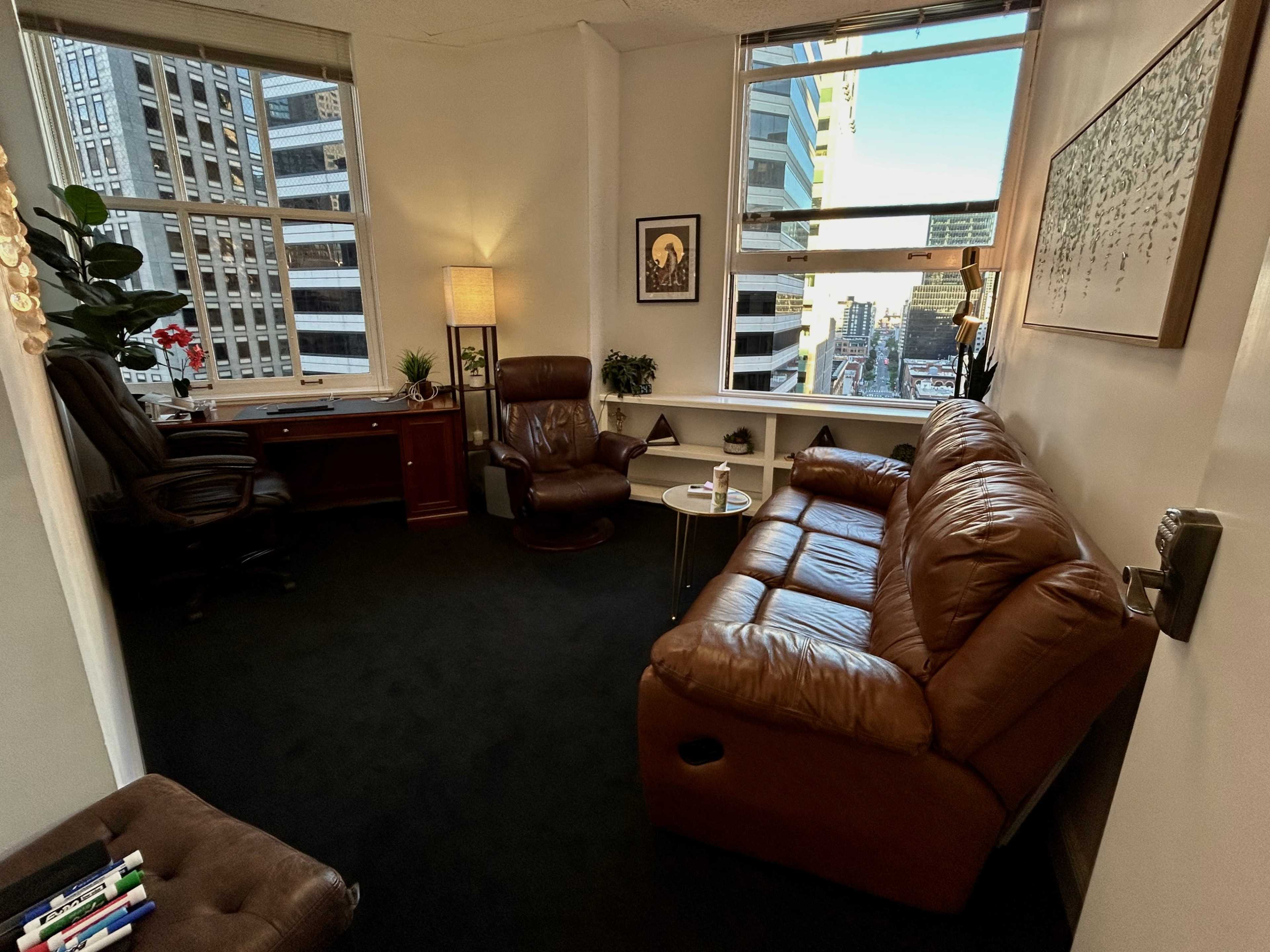A corner office with a brown leather sofa, an armchair, a desk with a computer, and large windows offering a view of a cityscape.