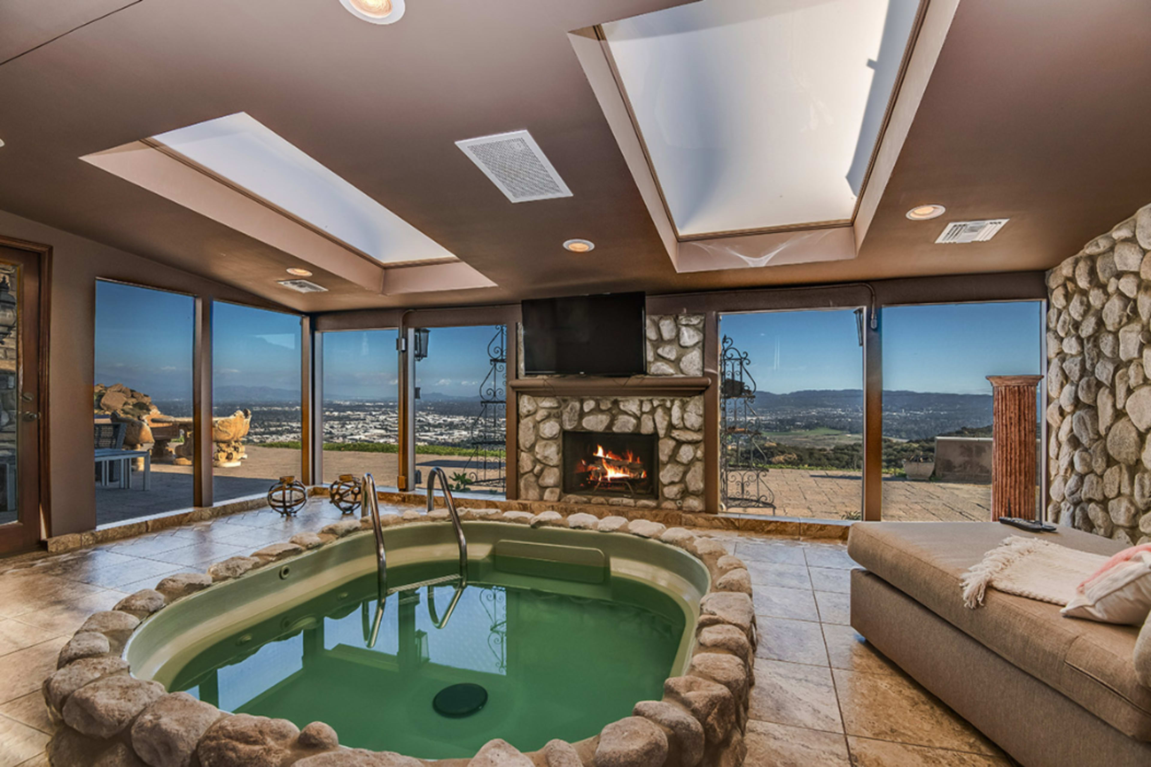 The image depicts a room featuring a circular stone-lined jacuzzi, a fireplace, large windows with scenic views, and a cozy seating area.