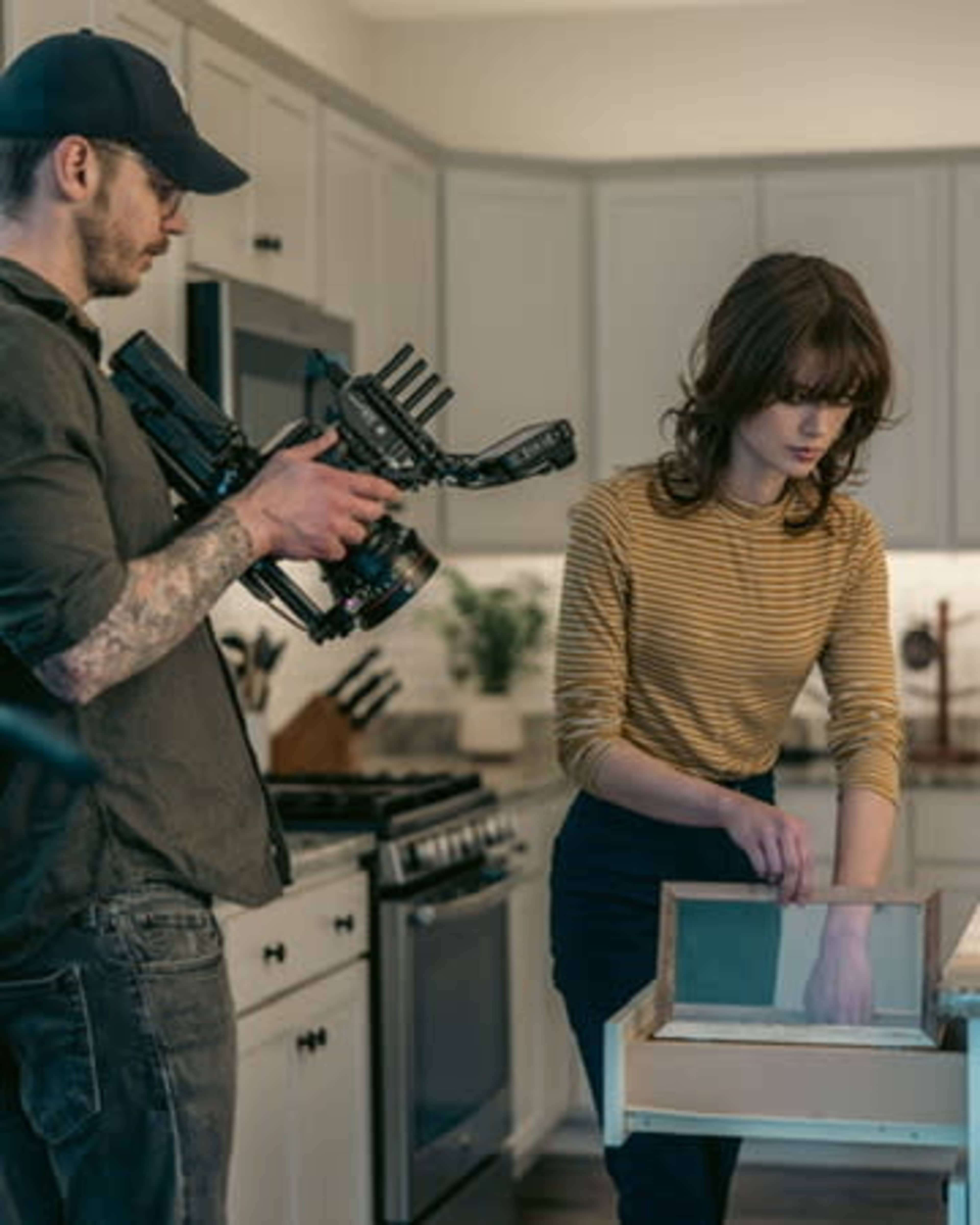 A man holds a camera while a woman organizes items in a drawer in a well-lit kitchen.