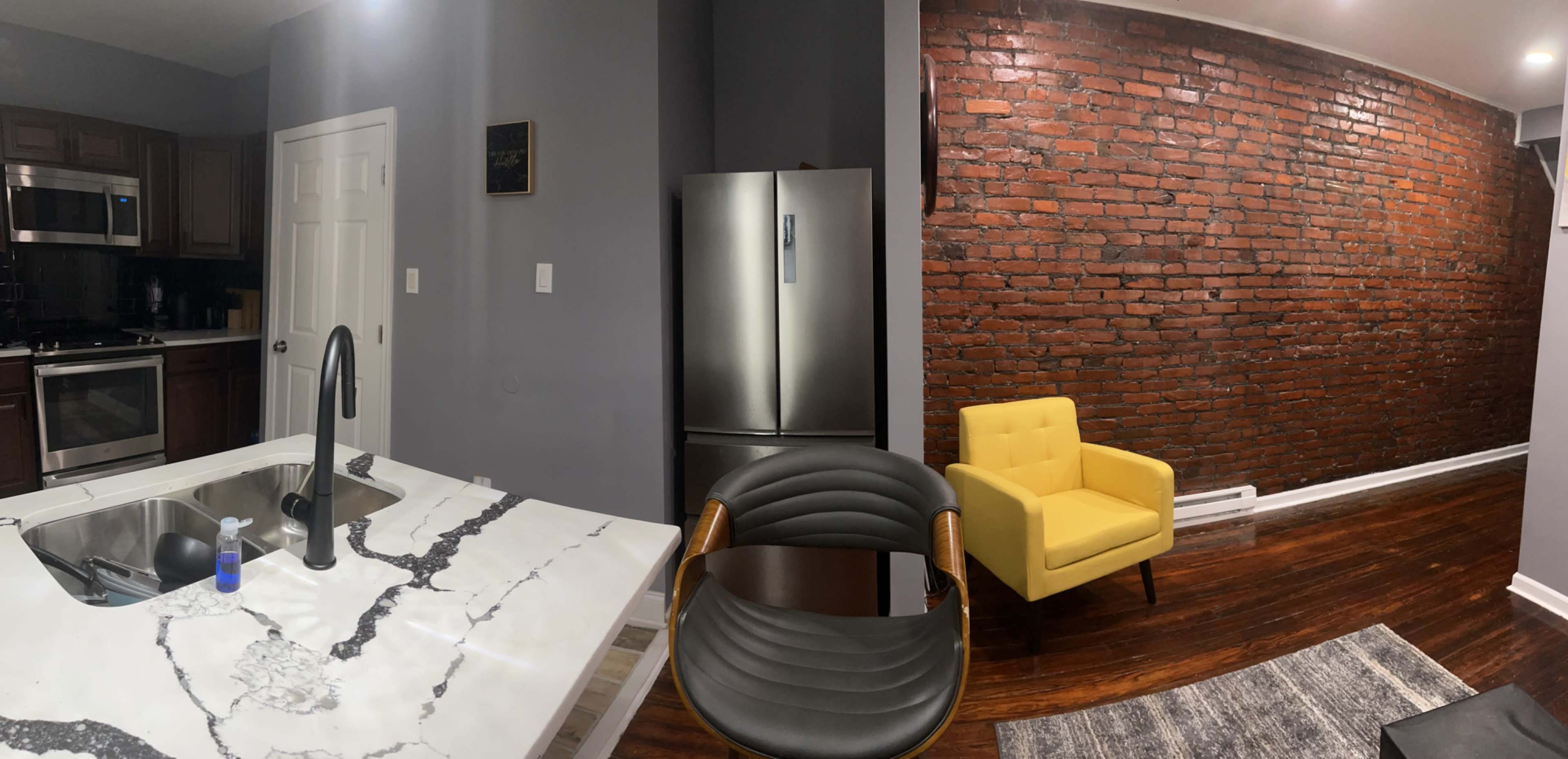 The image shows a modern kitchen with a marble counter, stainless steel refrigerator, and a yellow armchair against a brick wall.