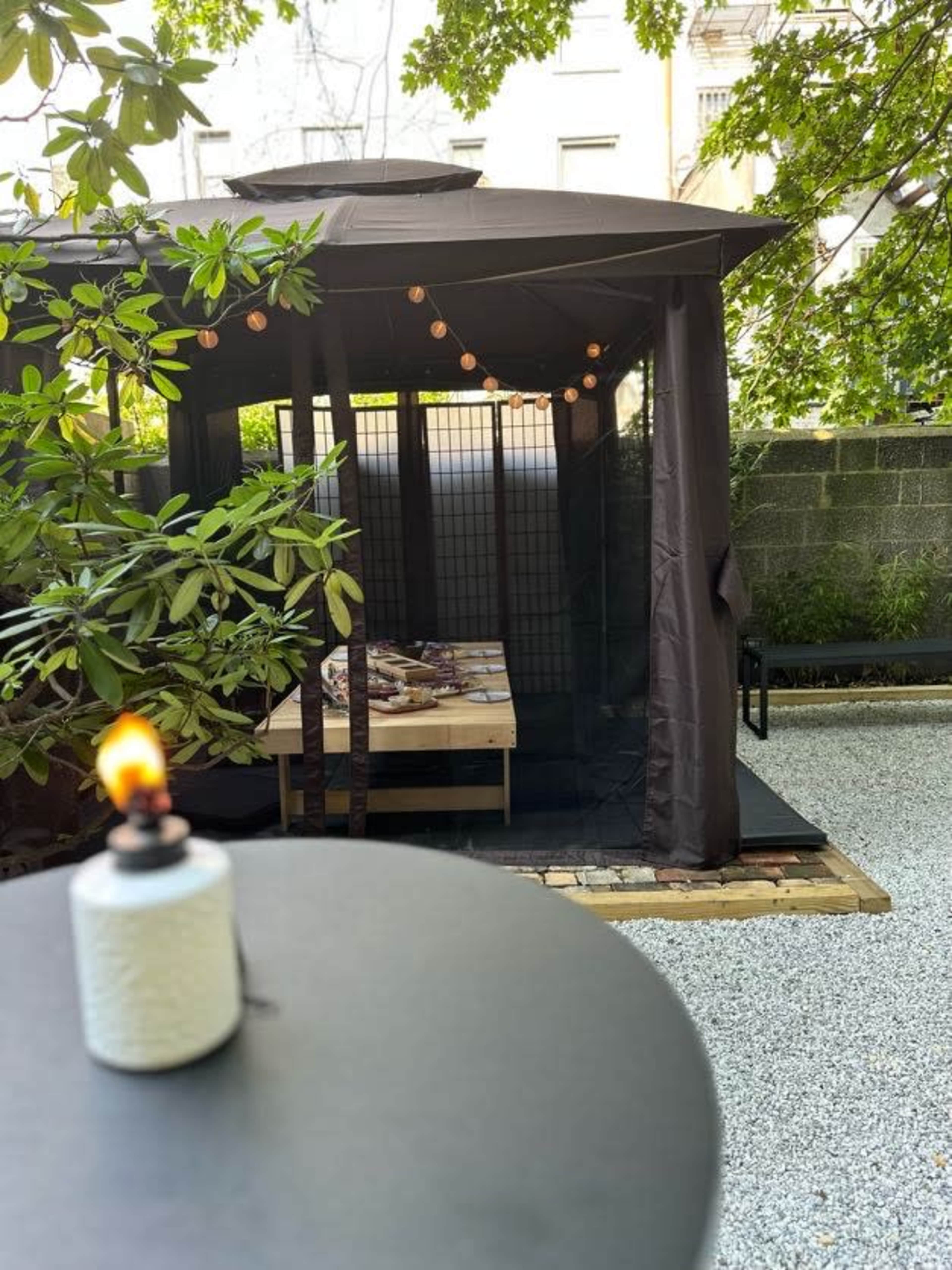 A dimly lit outdoor gazebo with string lights and a wooden platform is set within a landscaped area featuring gravel and greenery.