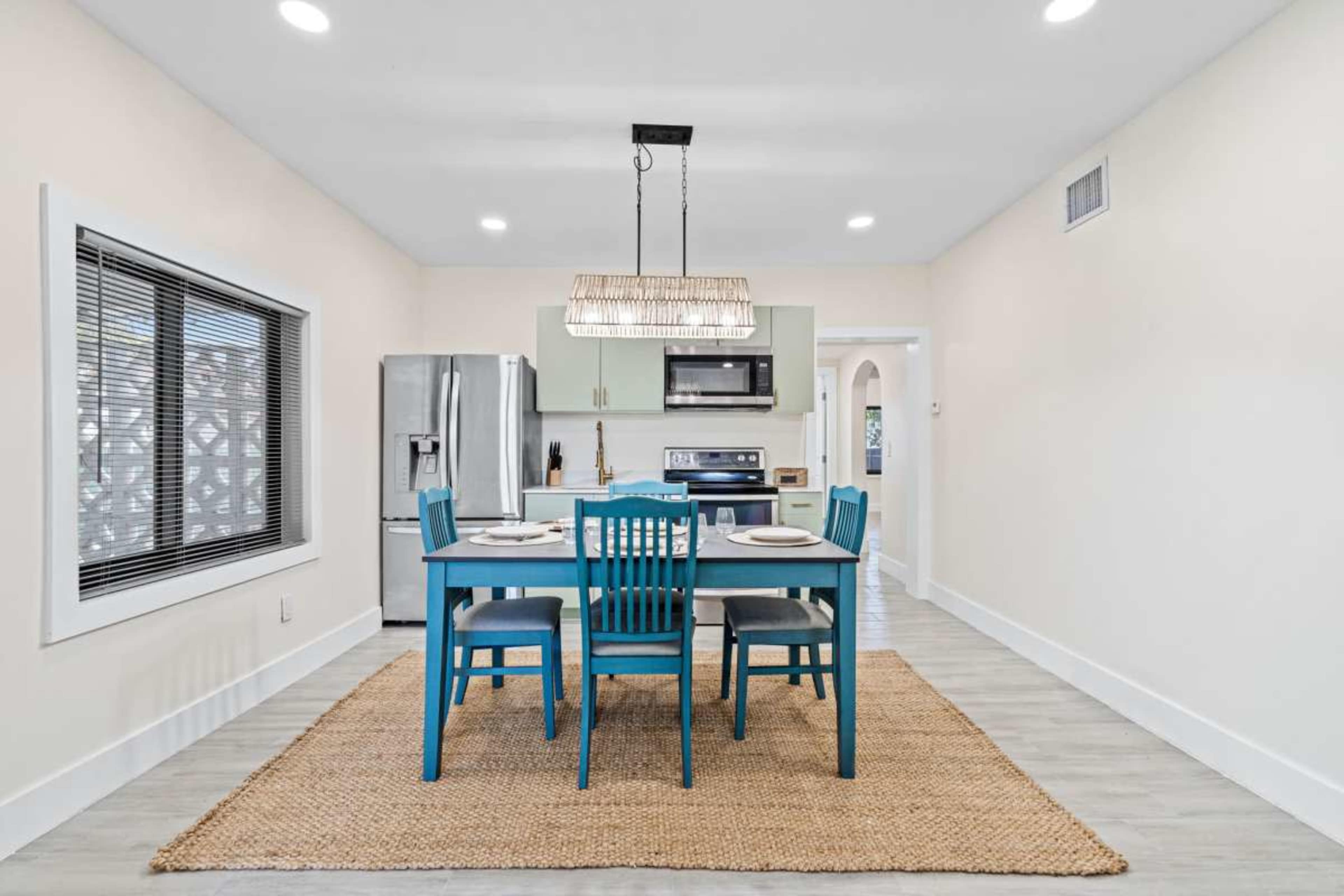 A bright kitchen features a blue dining table with chairs, a stainless steel refrigerator, and modern appliances against a light-colored backdrop.