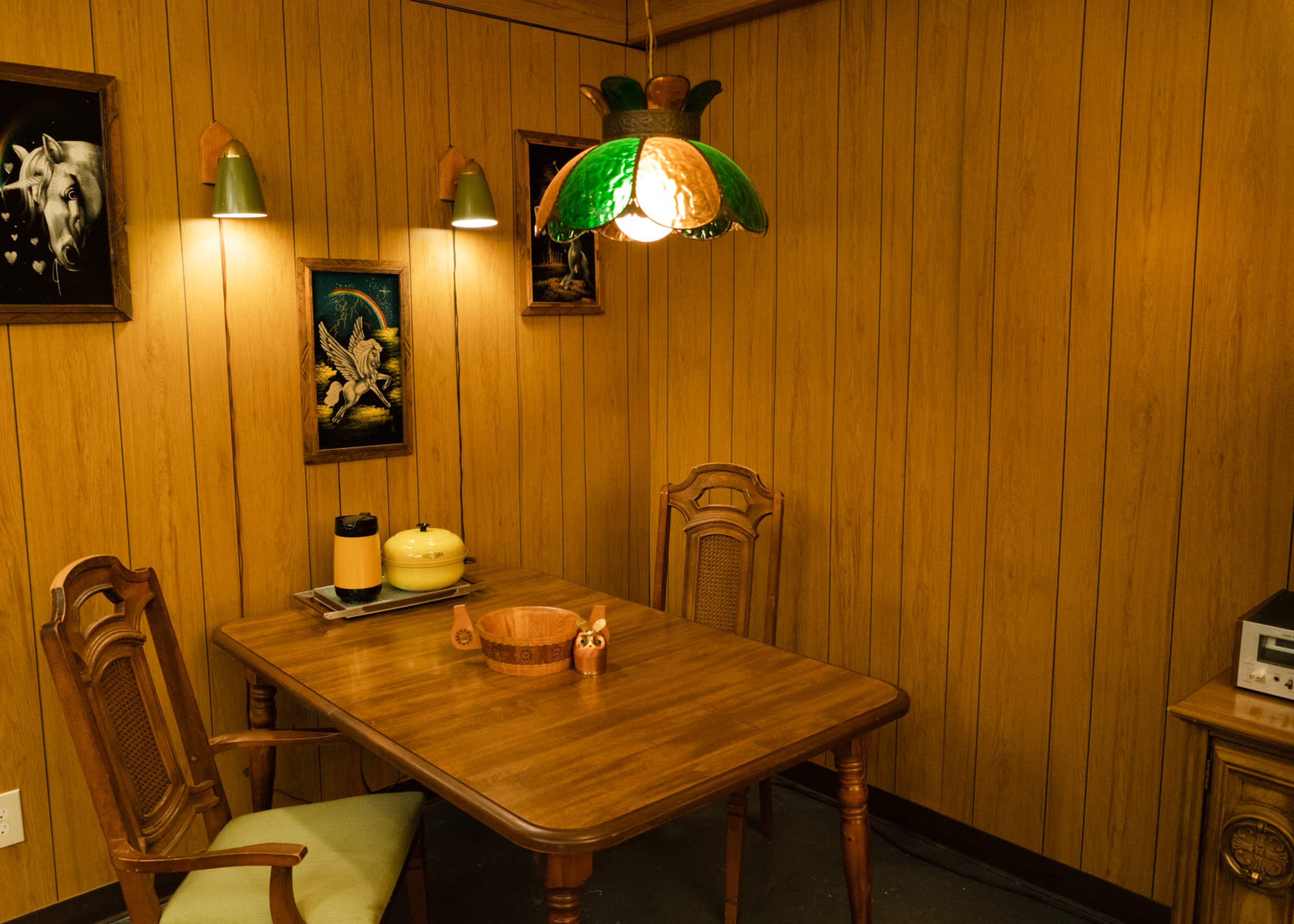 The image shows a wooden dining table with two chairs in a room featuring paneled walls and a green and orange pendant light.