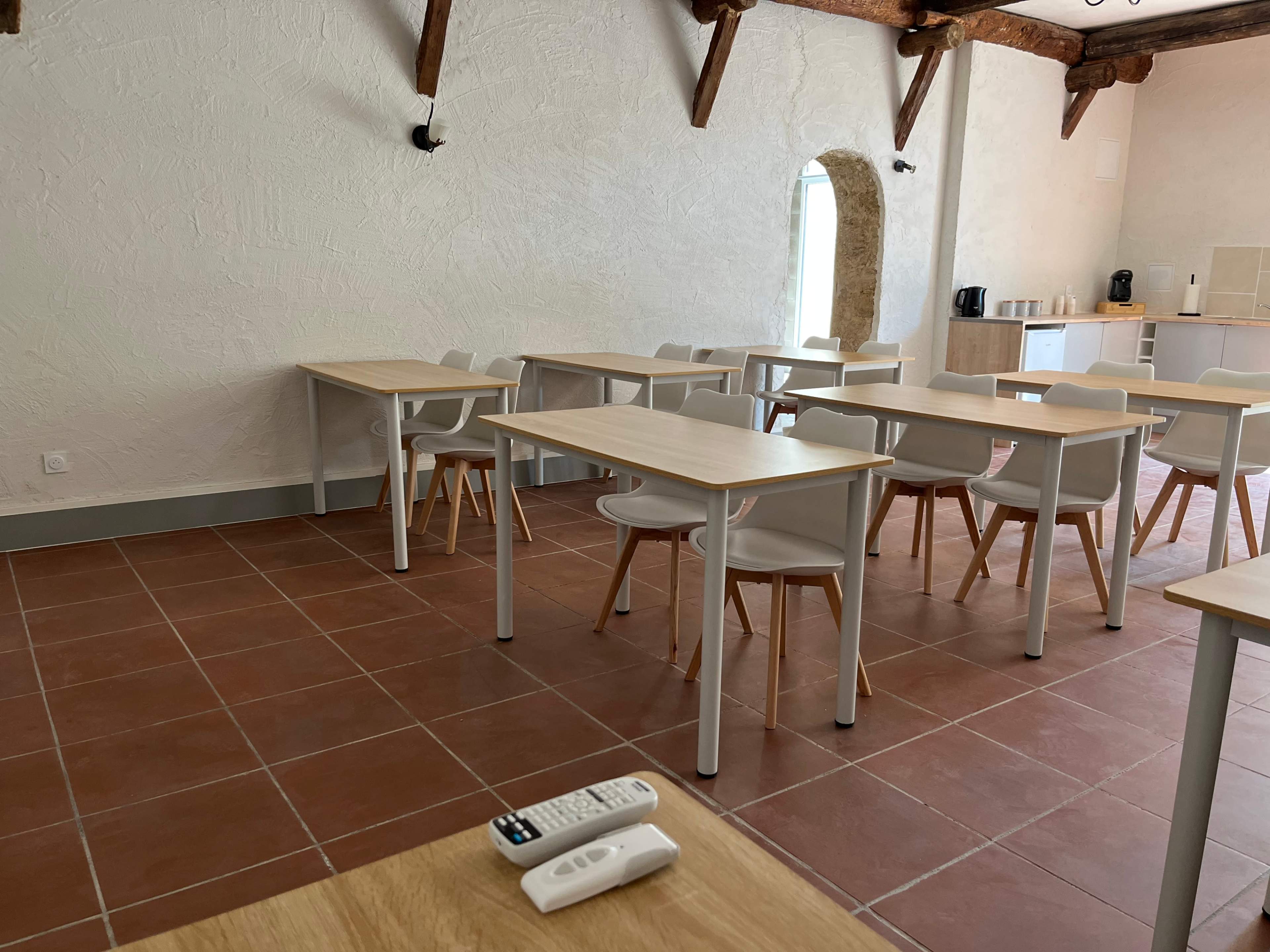 The image shows a nearly empty classroom with wooden tables and chairs arranged in a minimalist layout, set against a white textured wall and tiled floor.