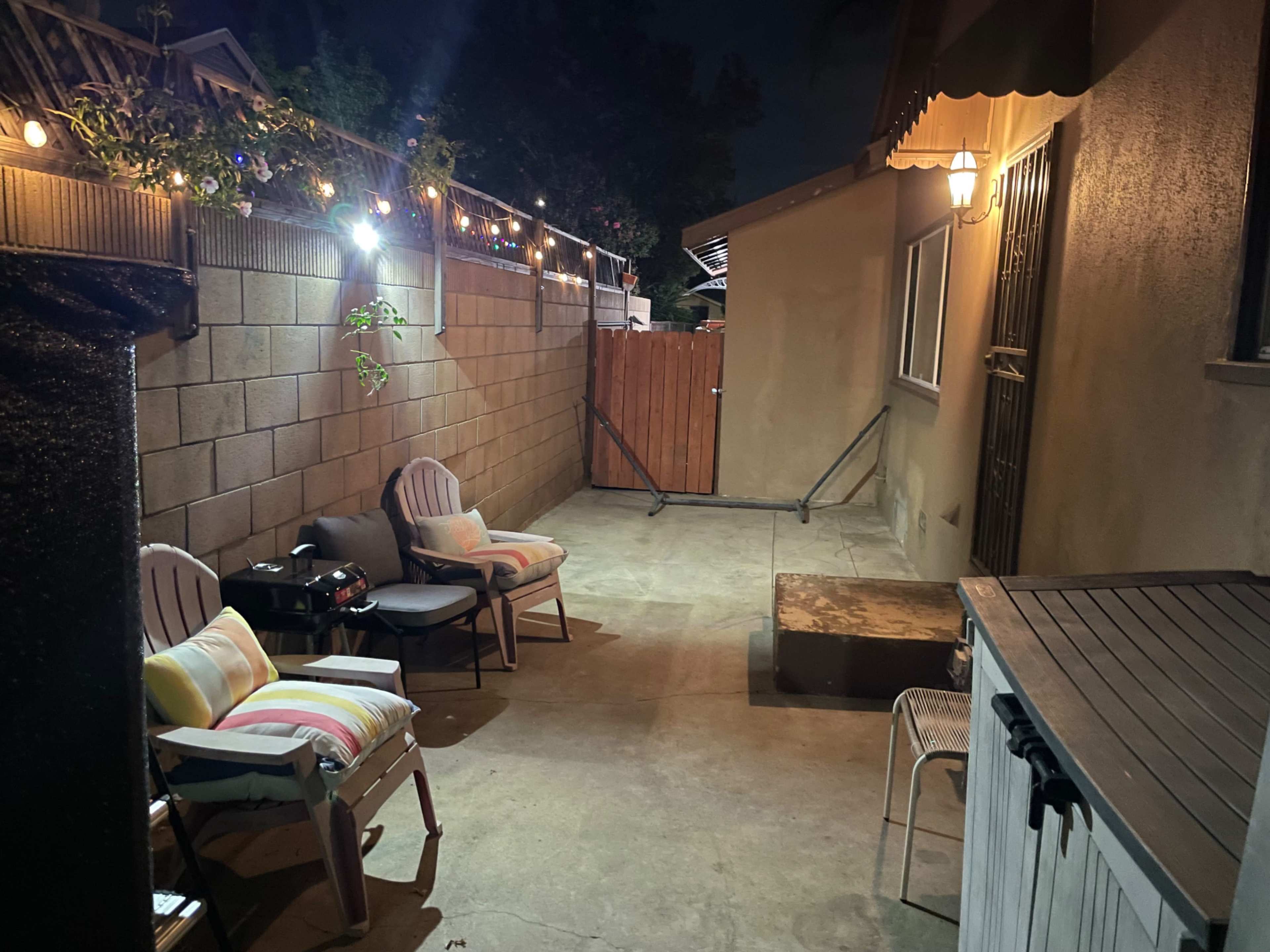 The image shows a small outdoor patio area at night, featuring a few chairs, string lights overhead, and a wall with a wooden gate.