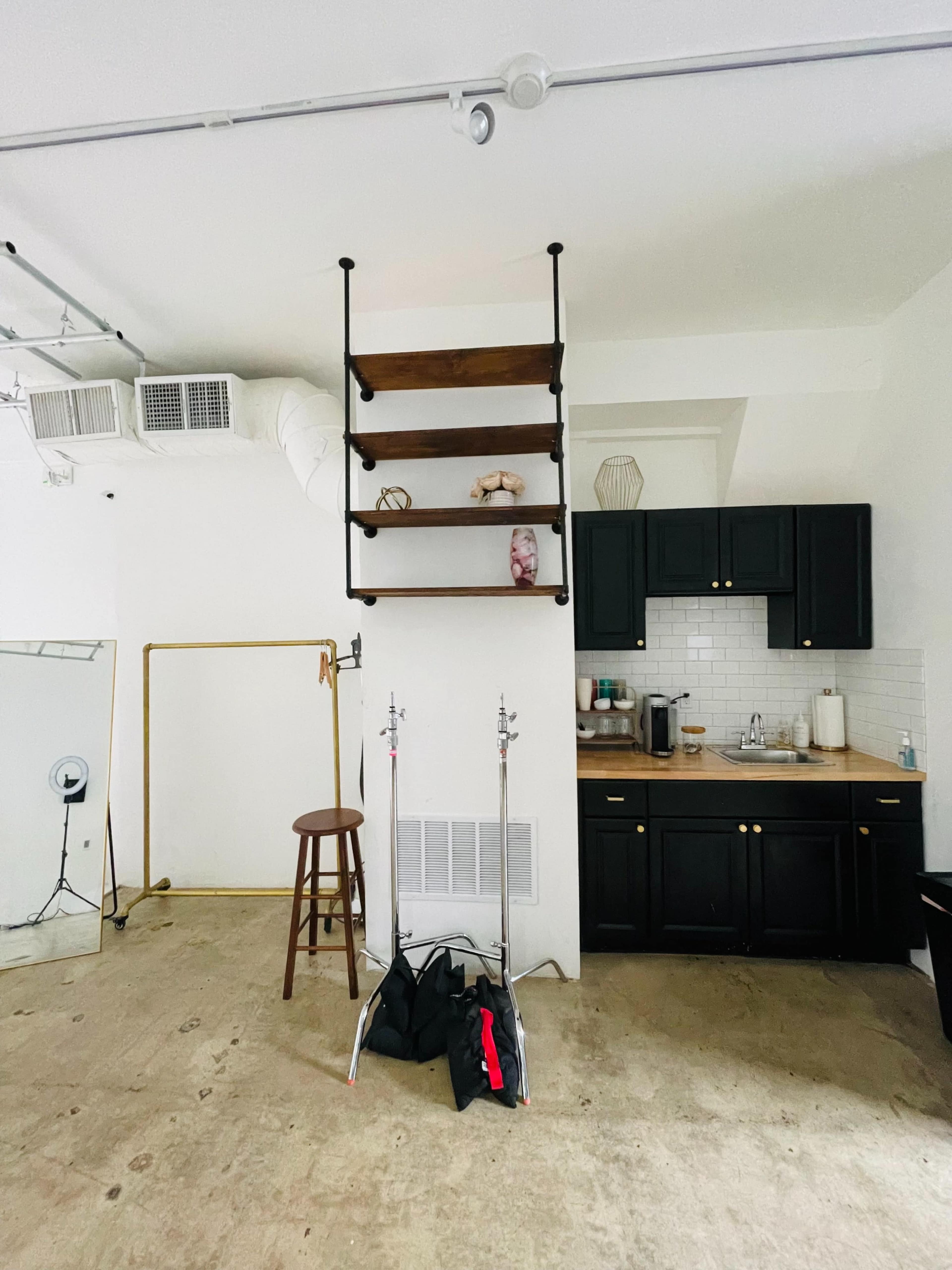 A partially furnished studio space with a kitchen area, a tall shelf against the wall, a bar stool, and some equipment on the floor.