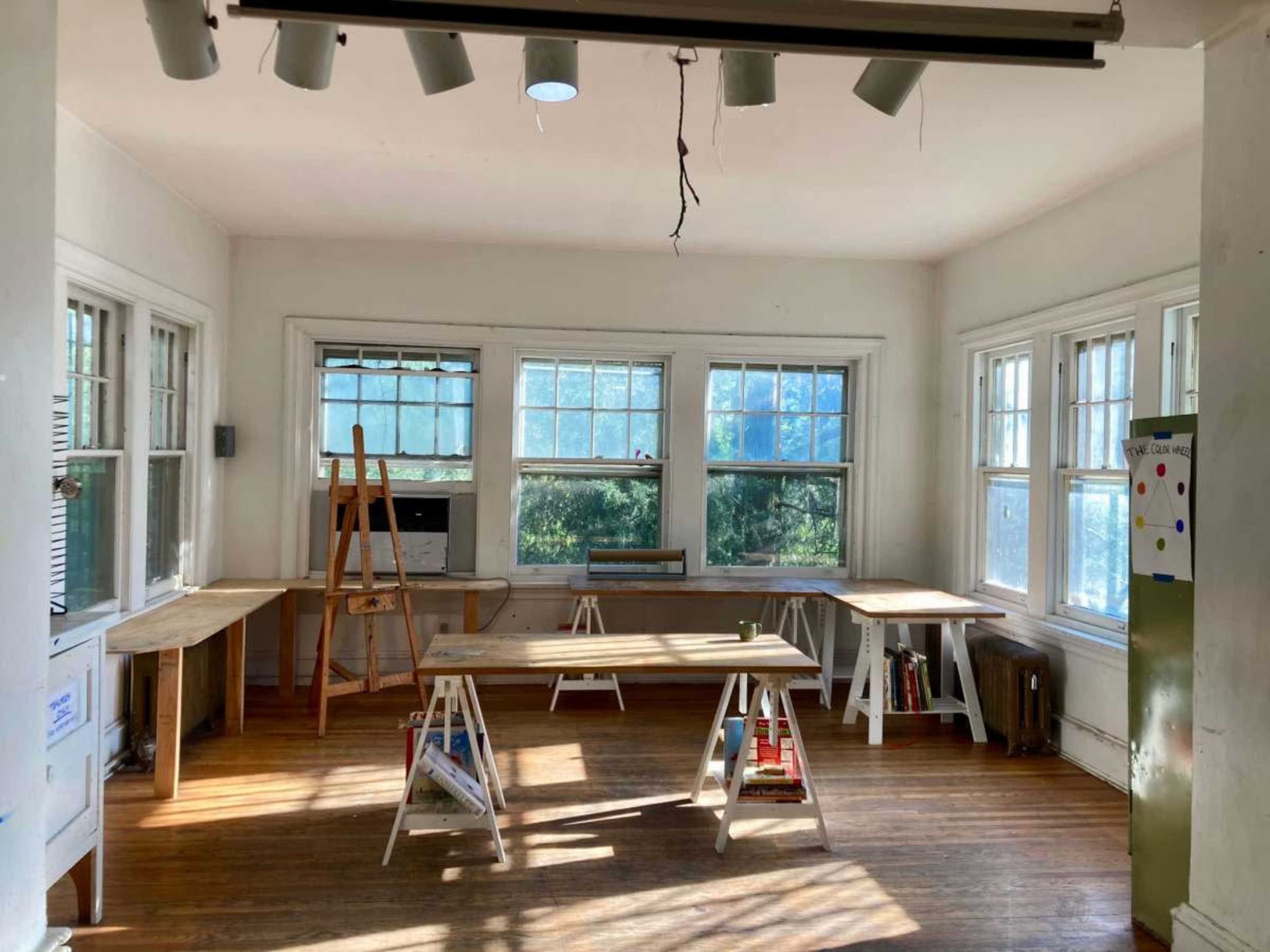 The image shows a bright, empty room with multiple wooden tables, large windows letting in natural light, and an easel in the corner.