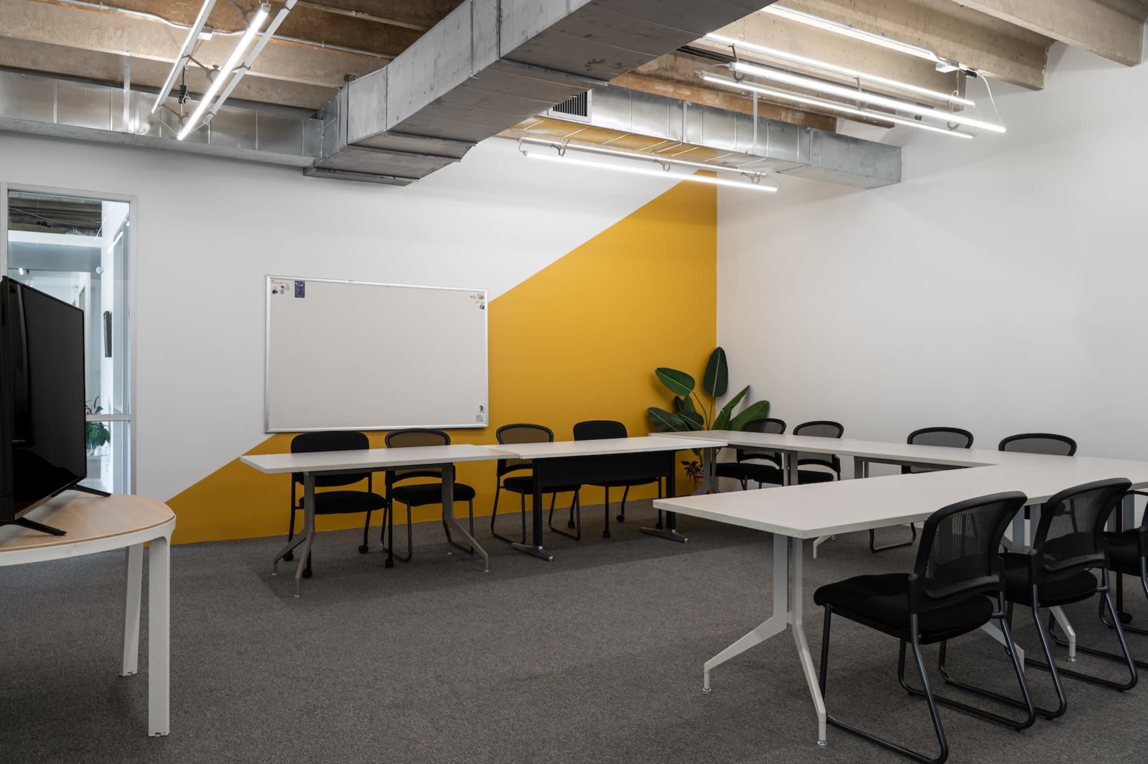 The image shows a modern meeting room with a whiteboard, a yellow accent wall, and several chairs arranged around a long table.
