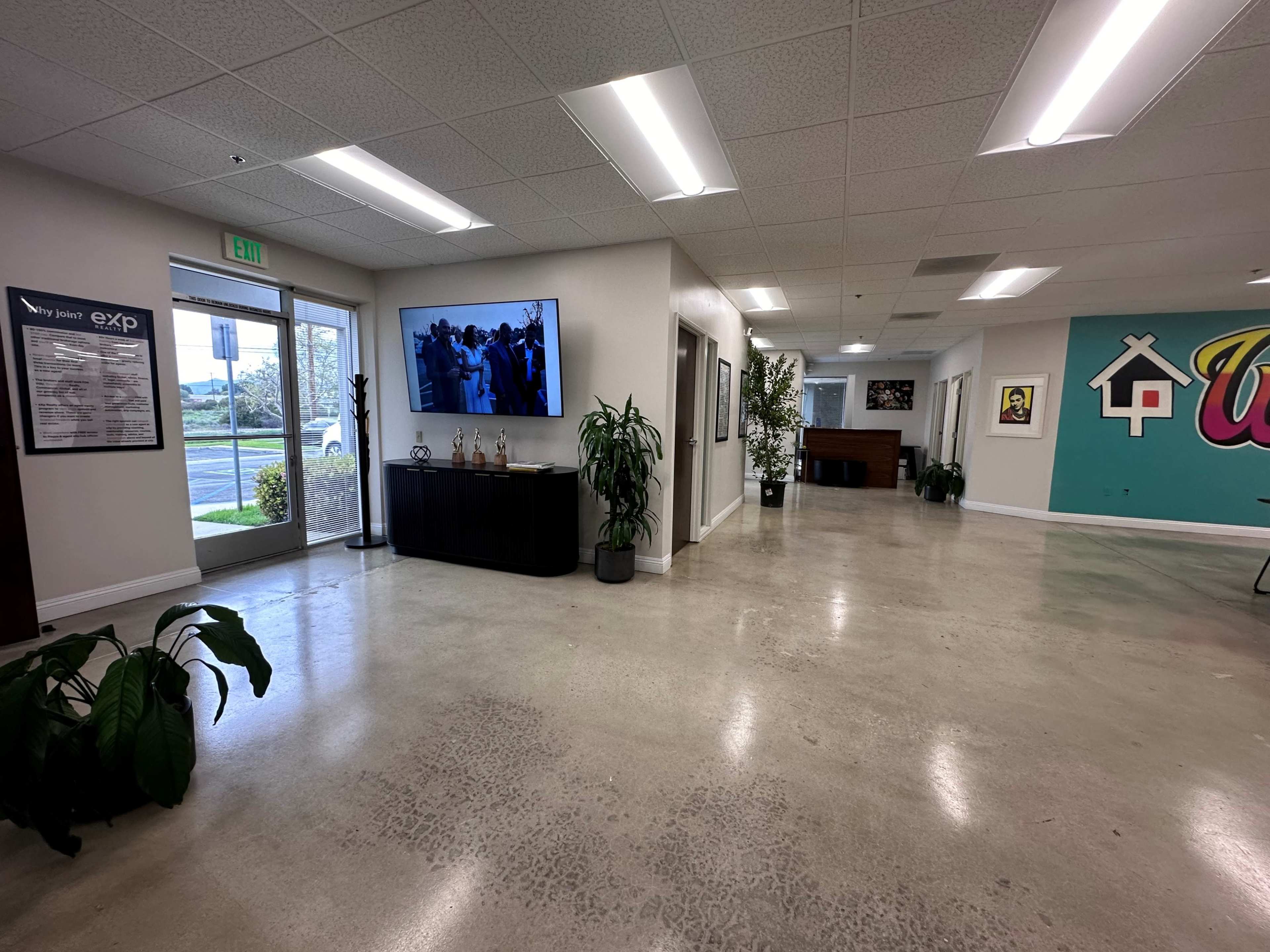 A spacious office interior with polished concrete floors, featuring a large TV mounted on the wall and plants scattered around.