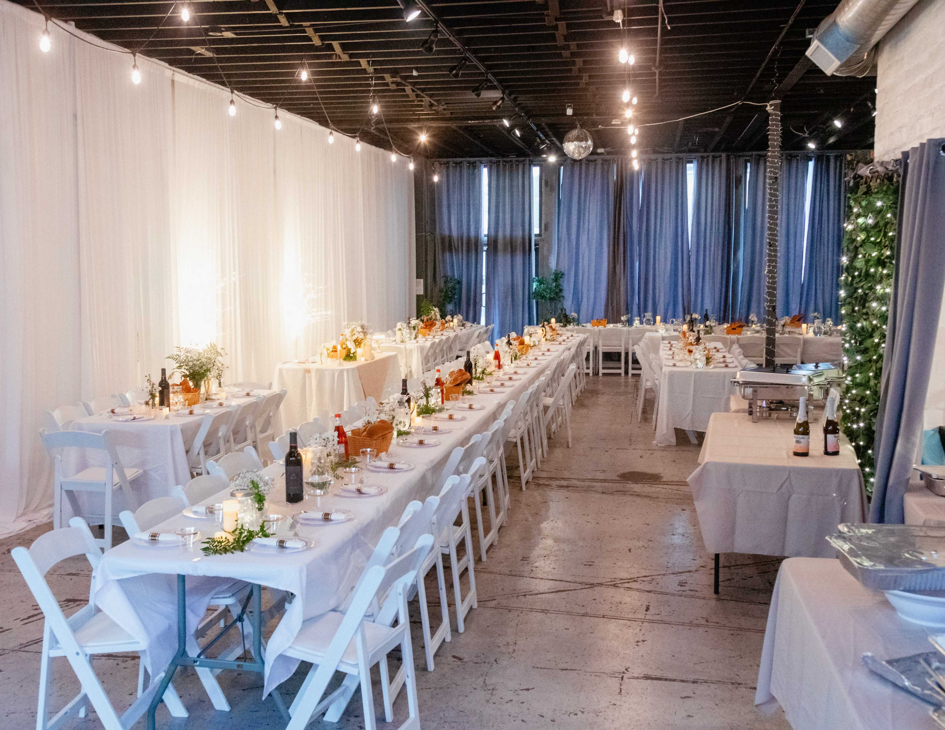 A spacious event venue is arranged with long tables set for a celebration, featuring decorations, food, and drinks under warm ambient lighting.