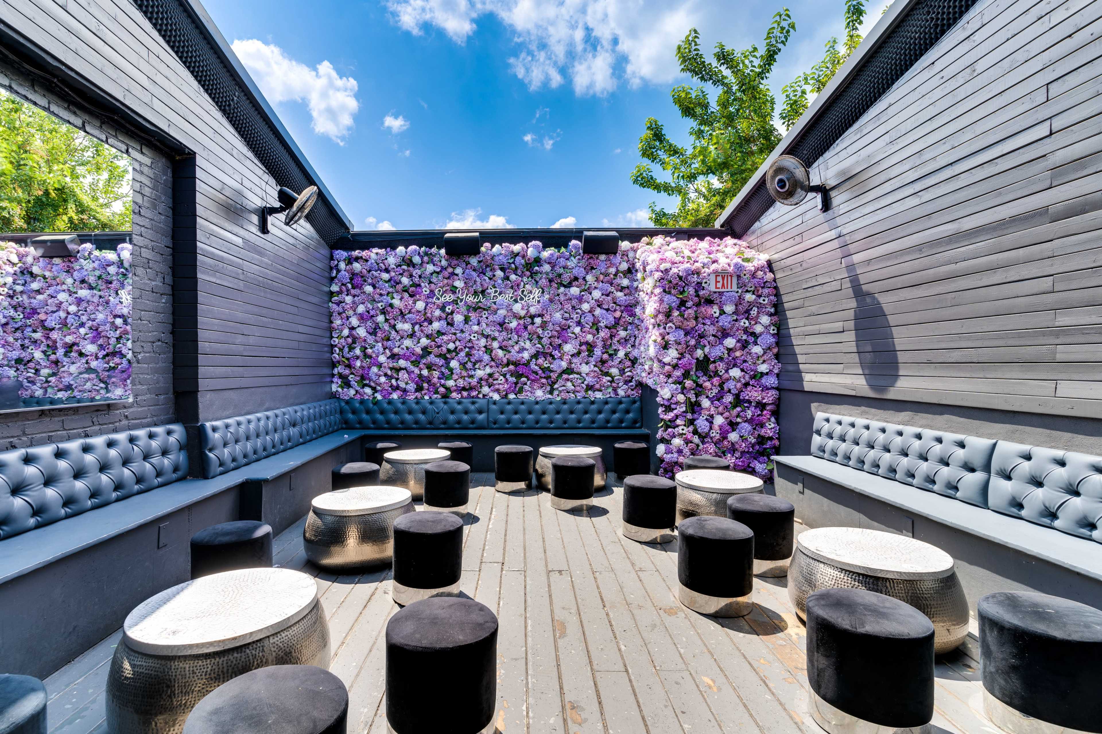 A stylish outdoor seating area features a wall of flowers and circular stools against a backdrop of blue sky and greenery.