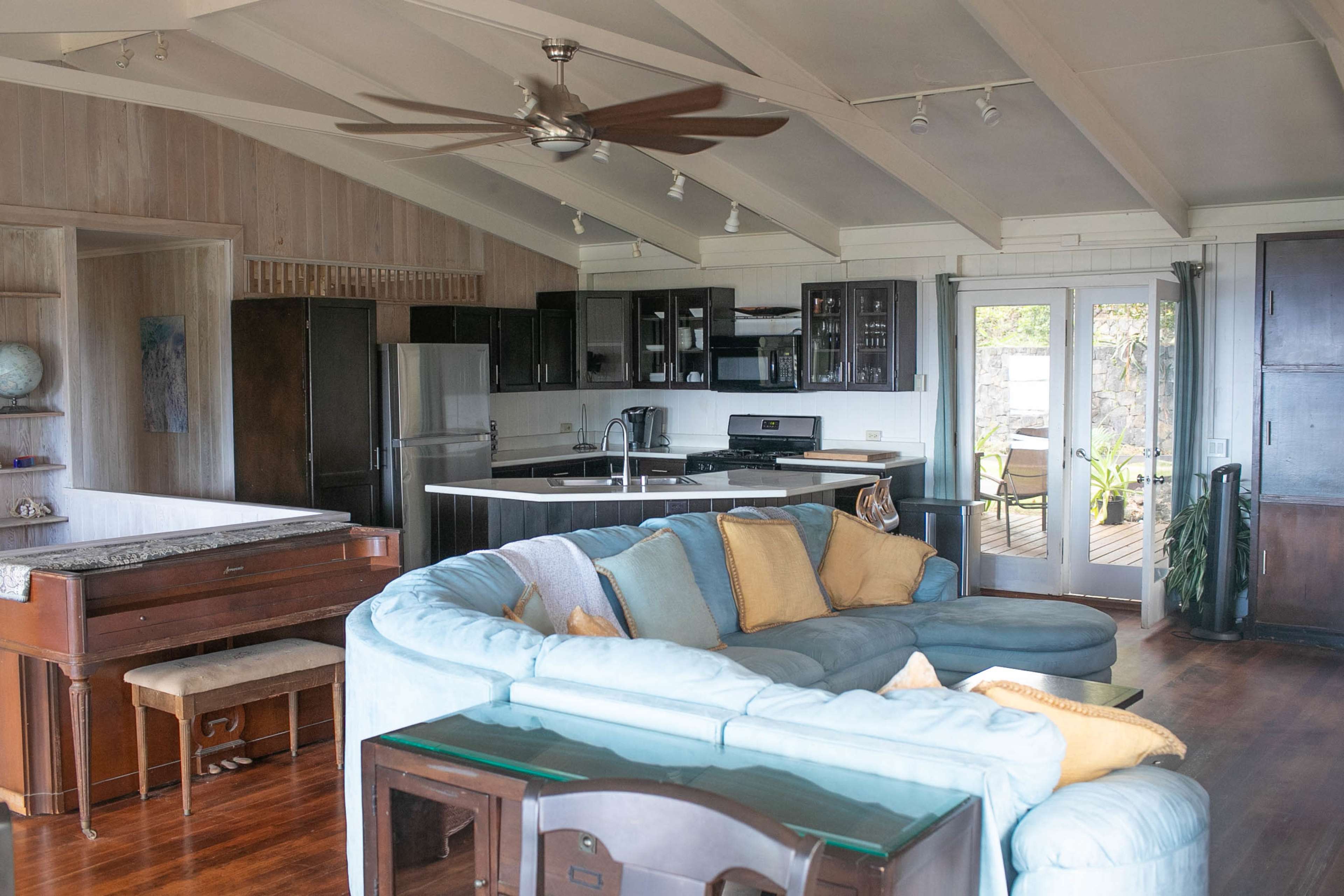 The interior of a modern coastal home features a spacious living area with a large curved sofa, a piano, and a kitchen with dark wood cabinetry.