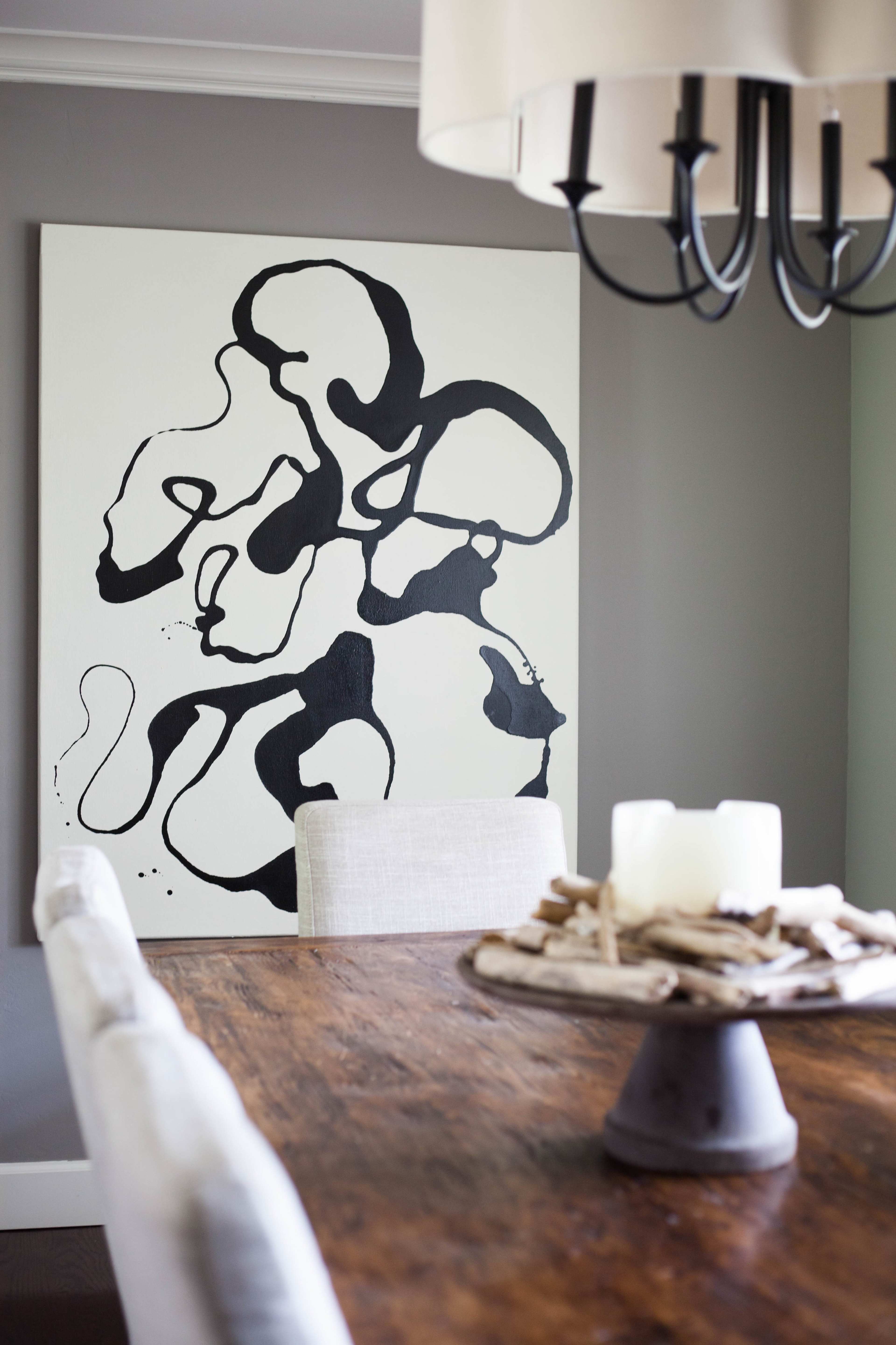 A dining table with a wooden surface is set in front of a large abstract black-and-white painting on a gray wall.