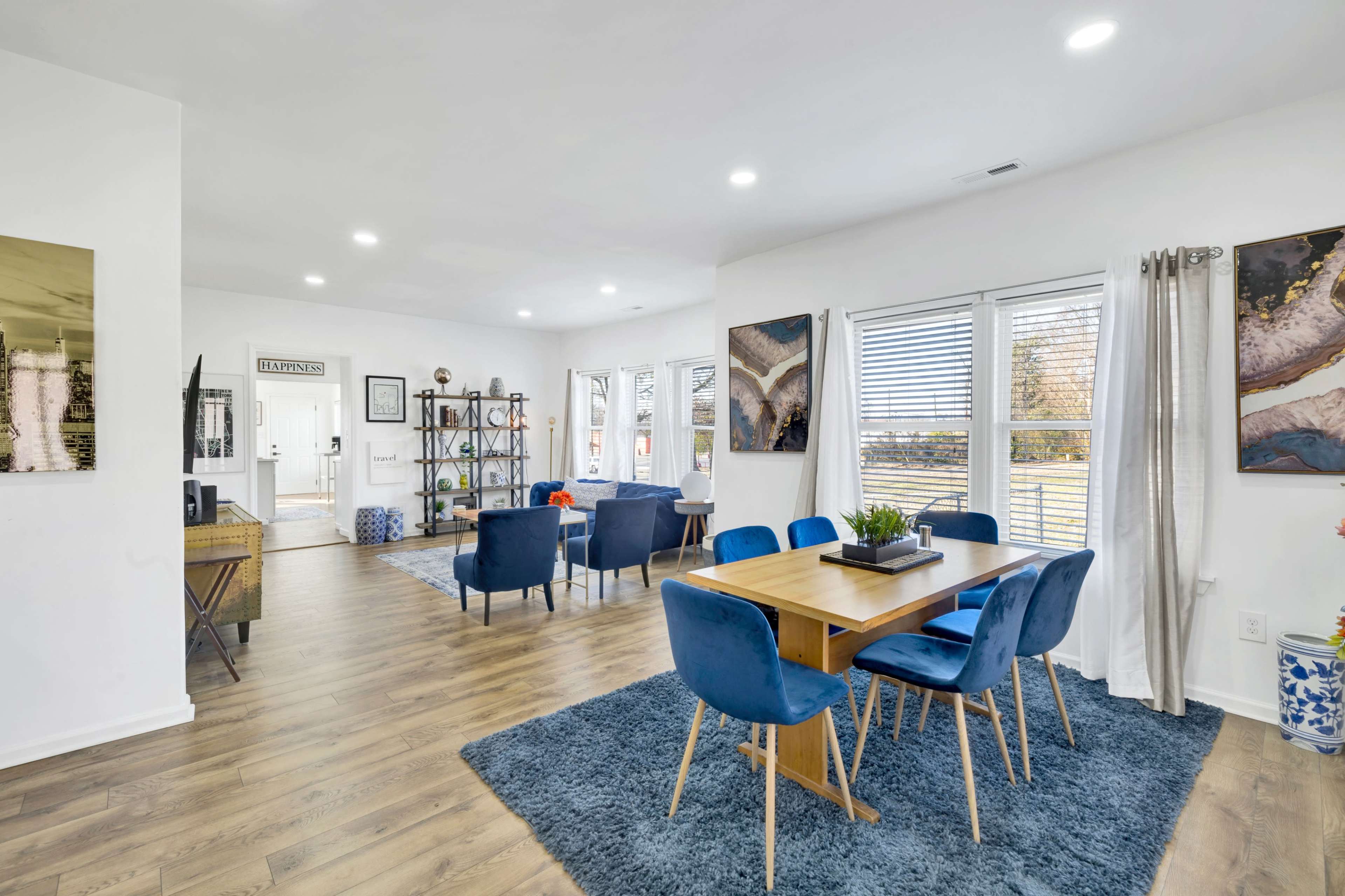 A bright, modern dining area features a wooden table surrounded by blue chairs, with large windows allowing natural light and a view of the outdoors.