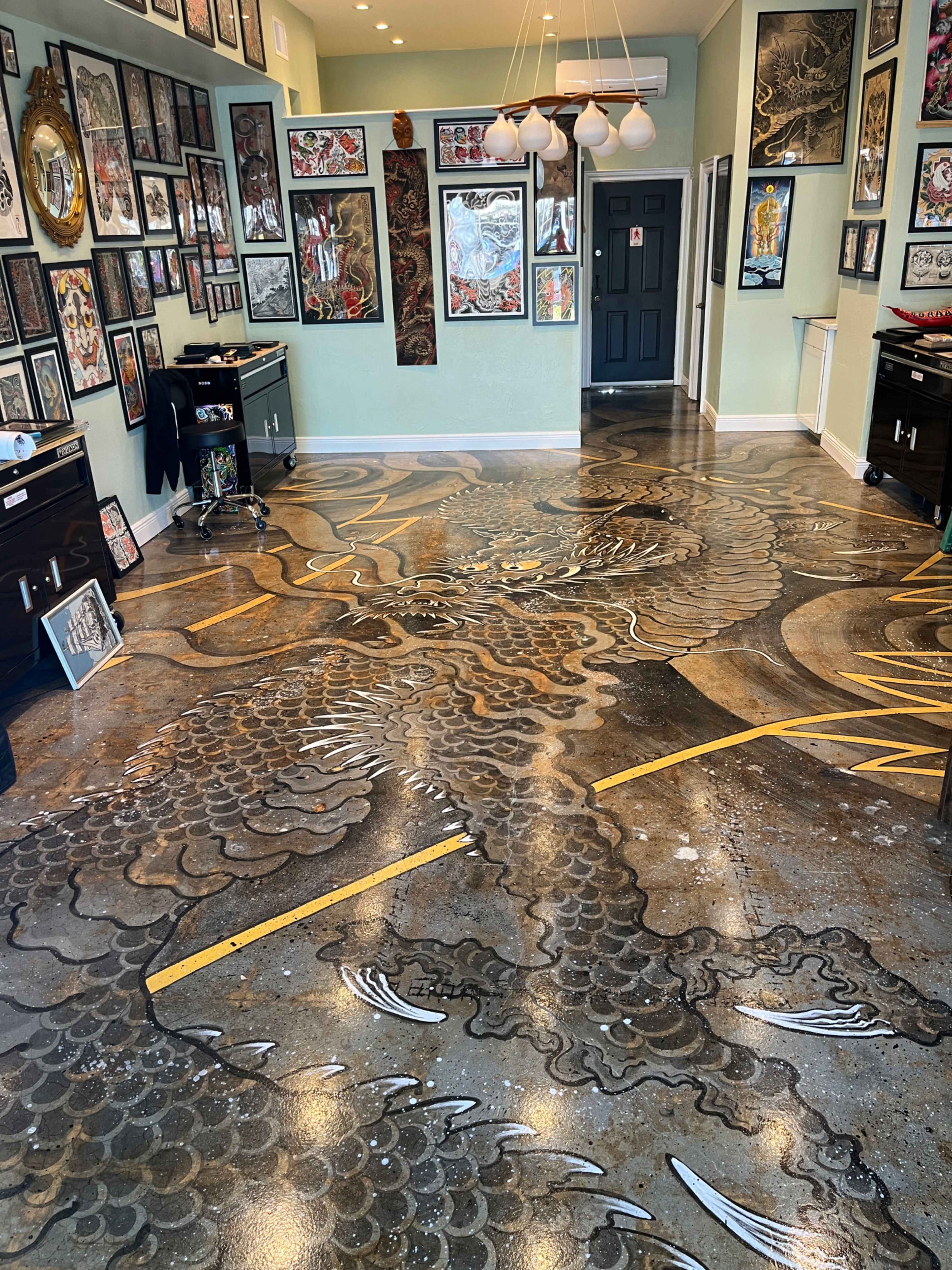 The image shows a tattoo studio with an intricately designed floor featuring a dragon motif, surrounded by numerous framed artworks on the walls.
