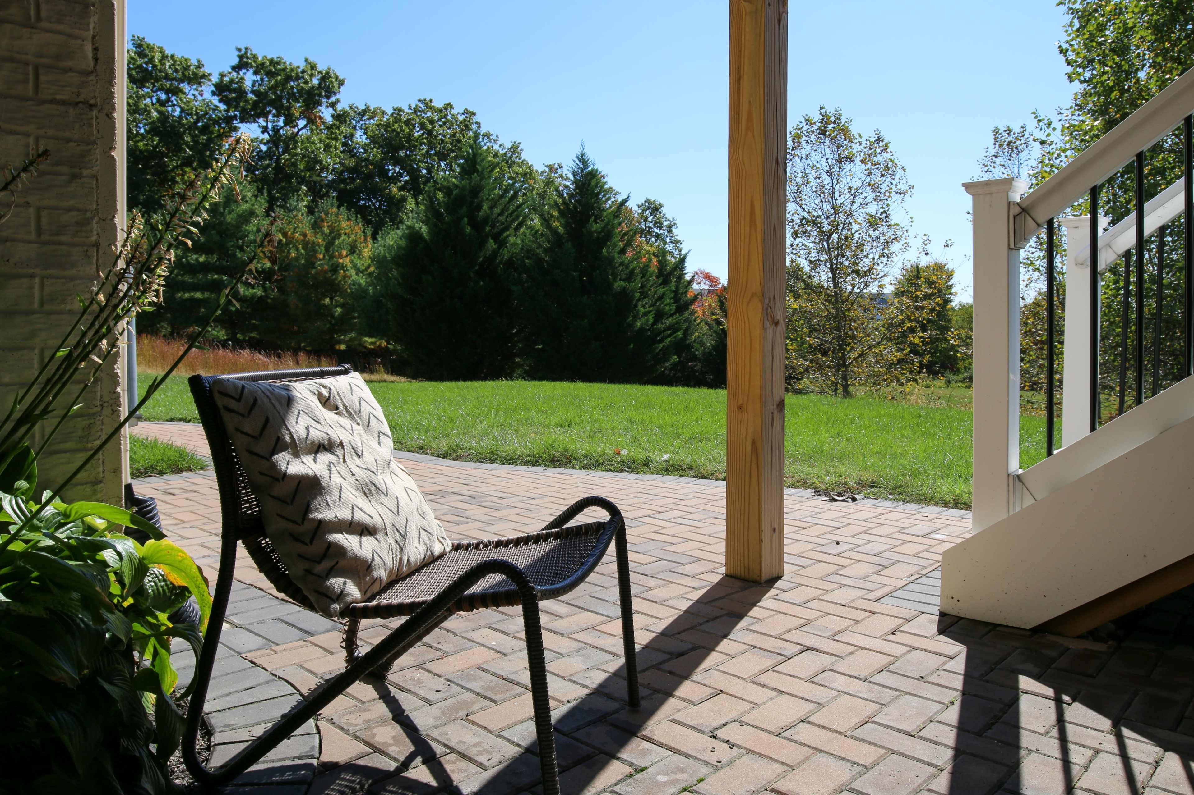 A chair with a patterned cushion is positioned on a brick patio, overlooking a grassy area and trees in the background under a clear sky.