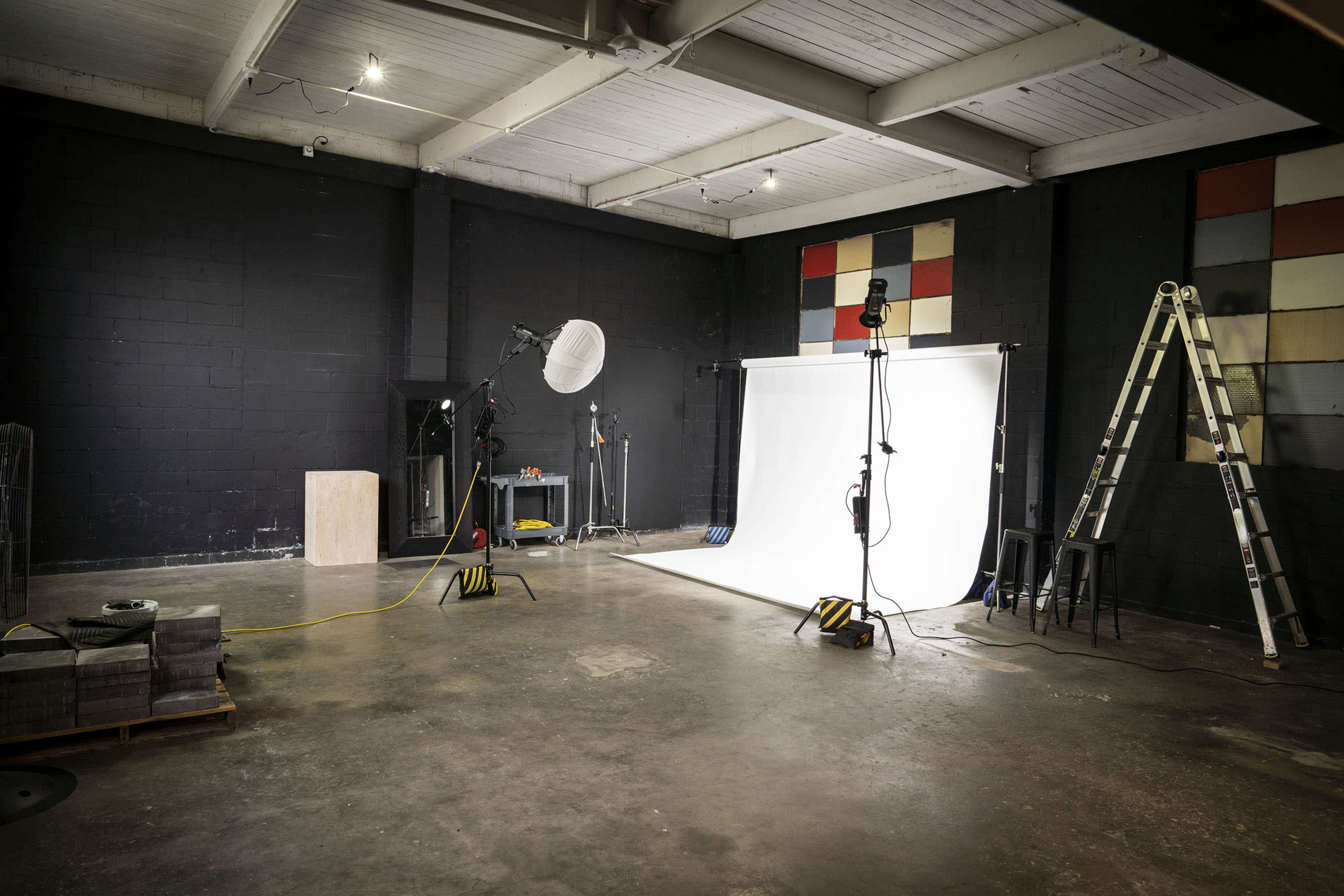 A photography studio with a white backdrop, lighting equipment, a ladder, and a variety of props scattered across the concrete floor.