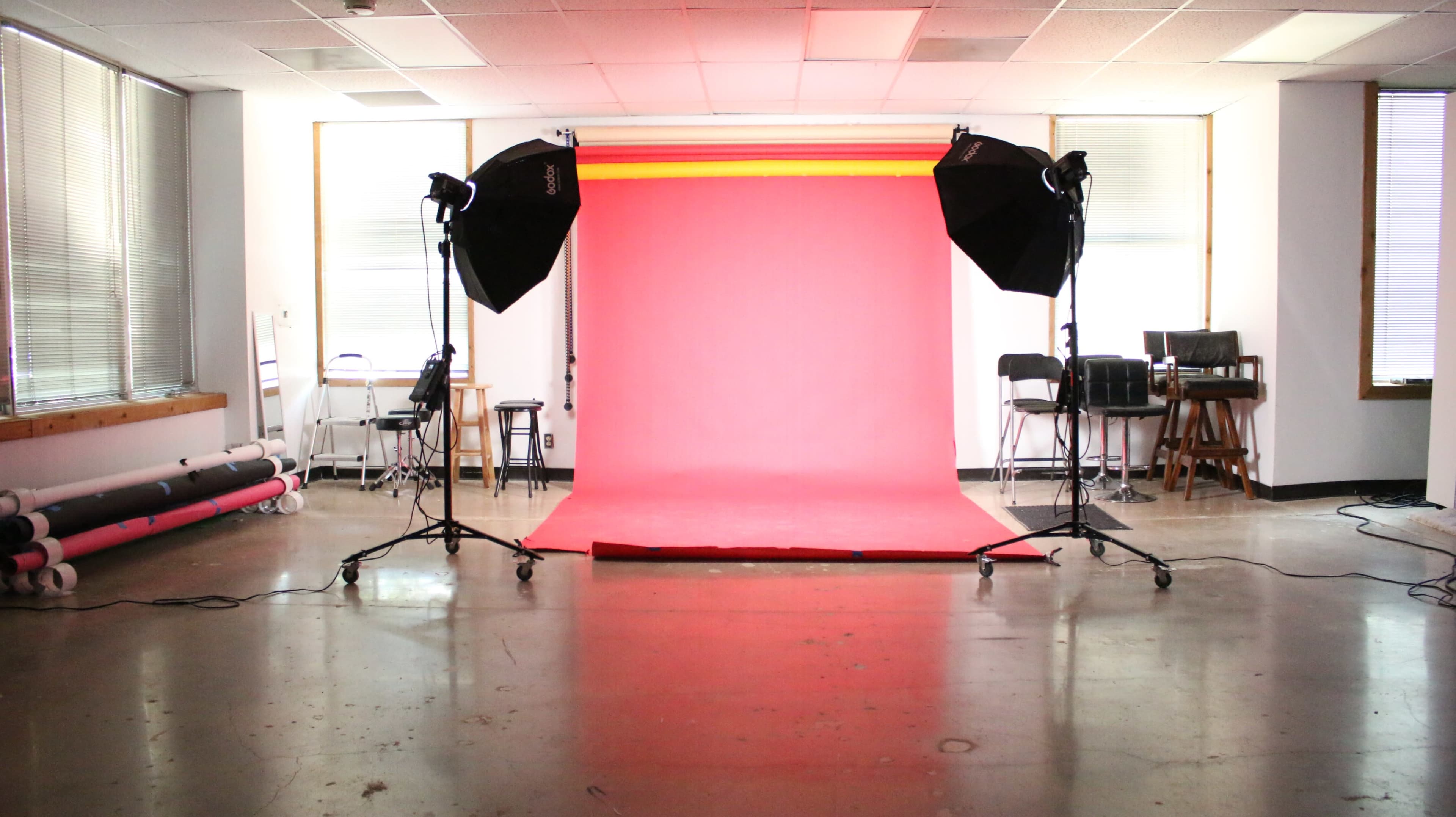 A photography studio features a pink backdrop with two studio lights and chairs arranged along the sides.