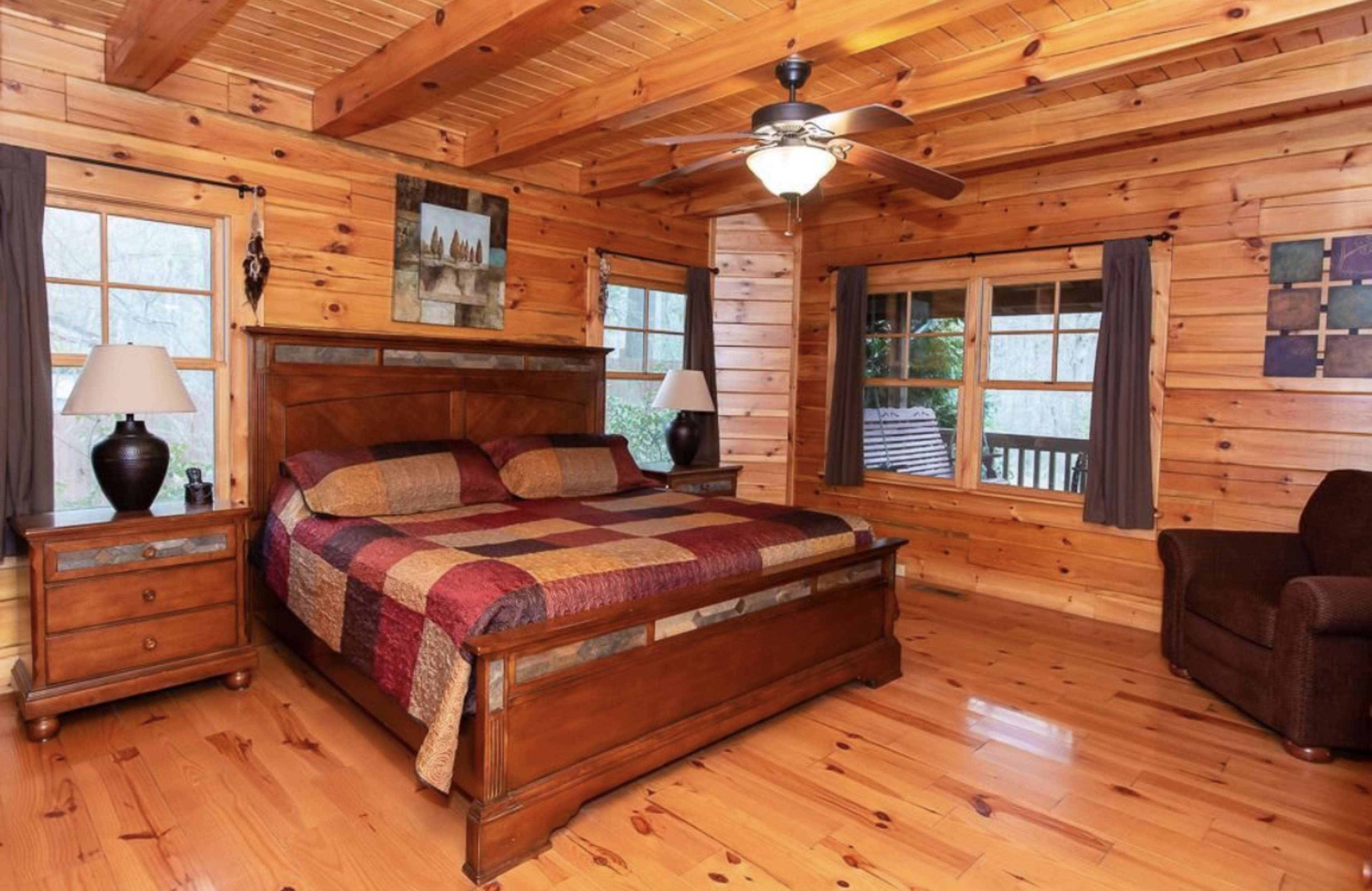 The image shows a cozy wooden bedroom with a large bed, two nightstands, and windows that overlook a wooded area.