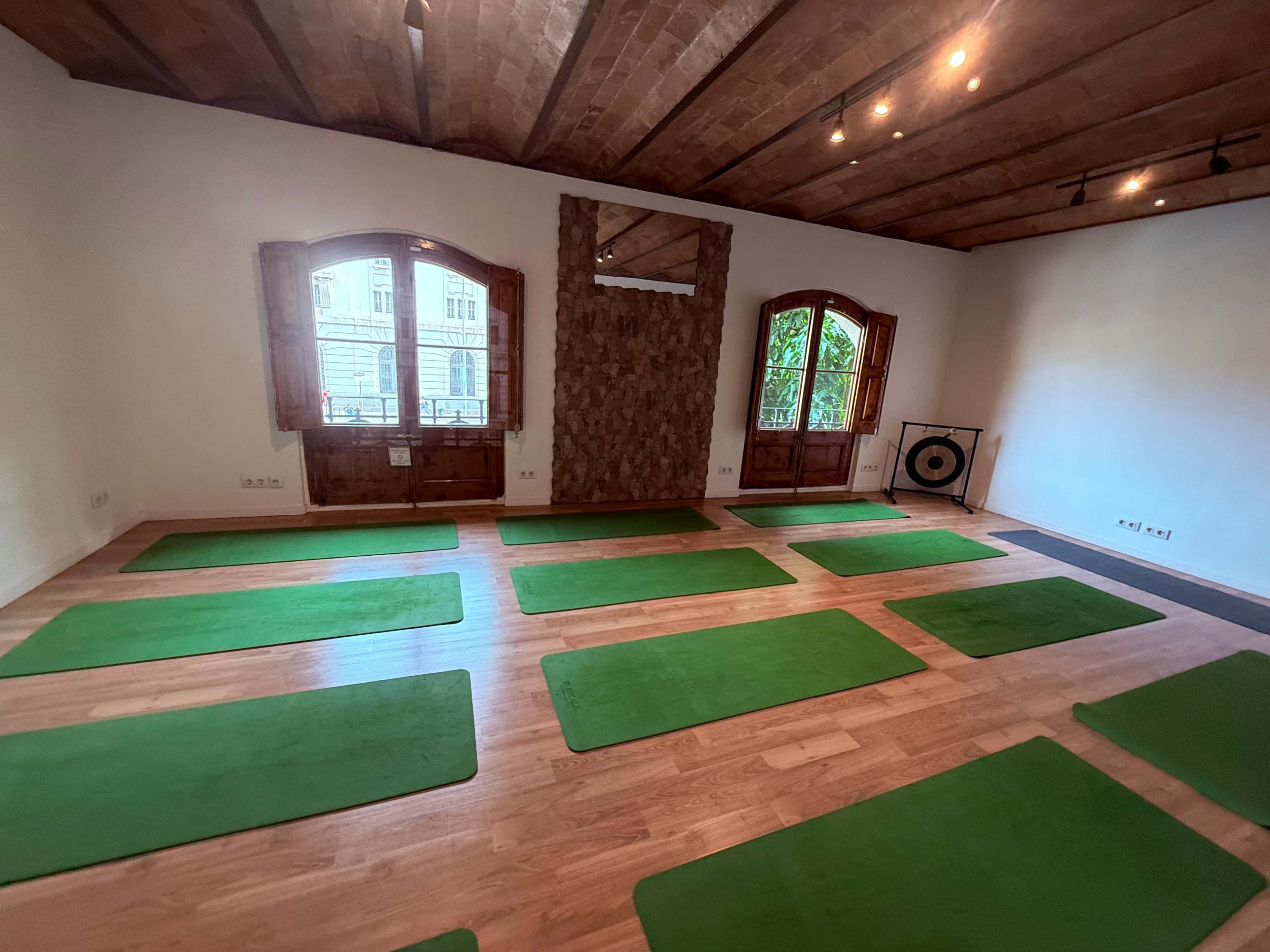 The image shows a spacious yoga studio with green mats arranged on a wooden floor, illuminated by natural light from large windows.