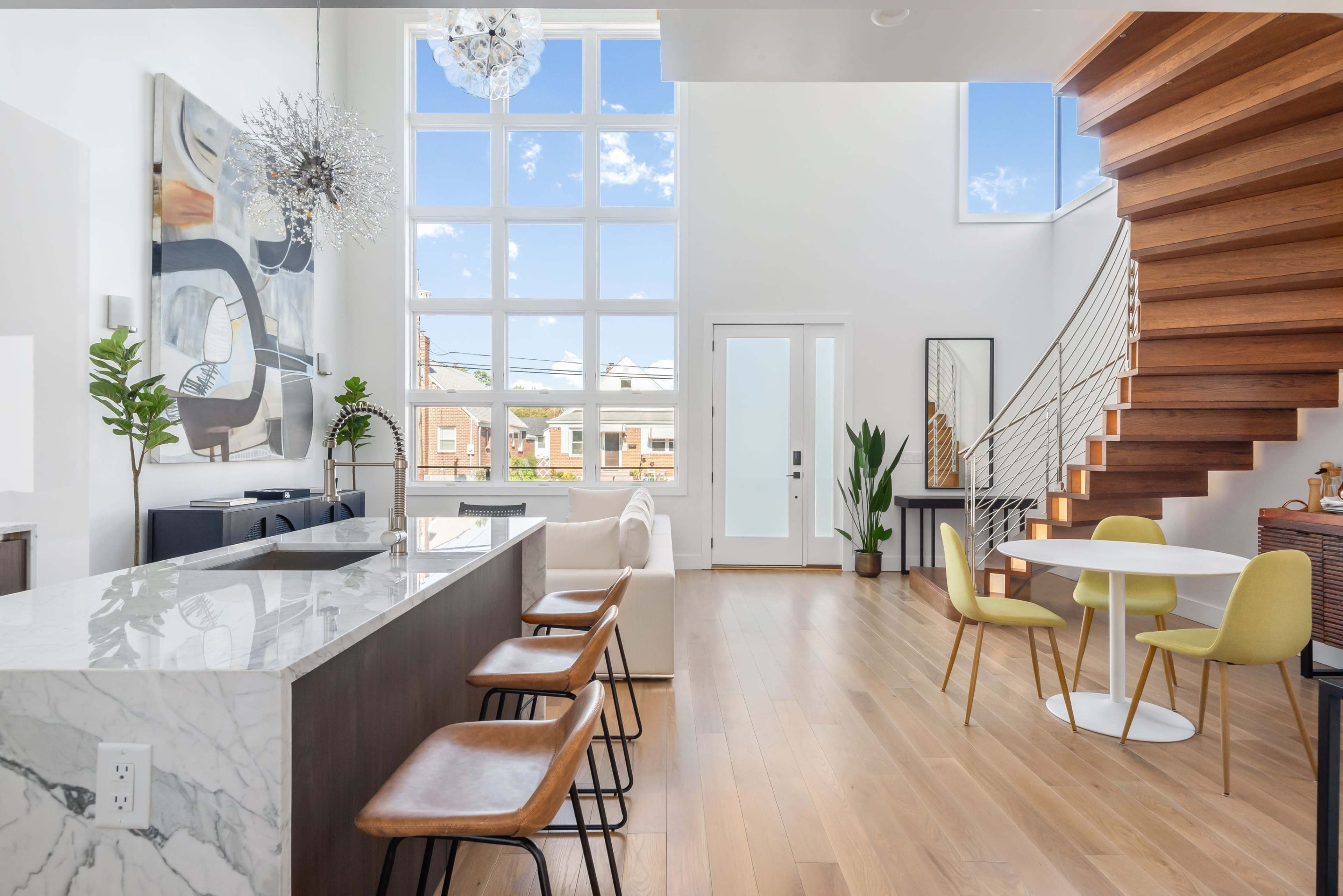 A modern interior with a marble kitchen island, a wooden staircase, large windows, and a dining area featuring a round table and colorful chairs.