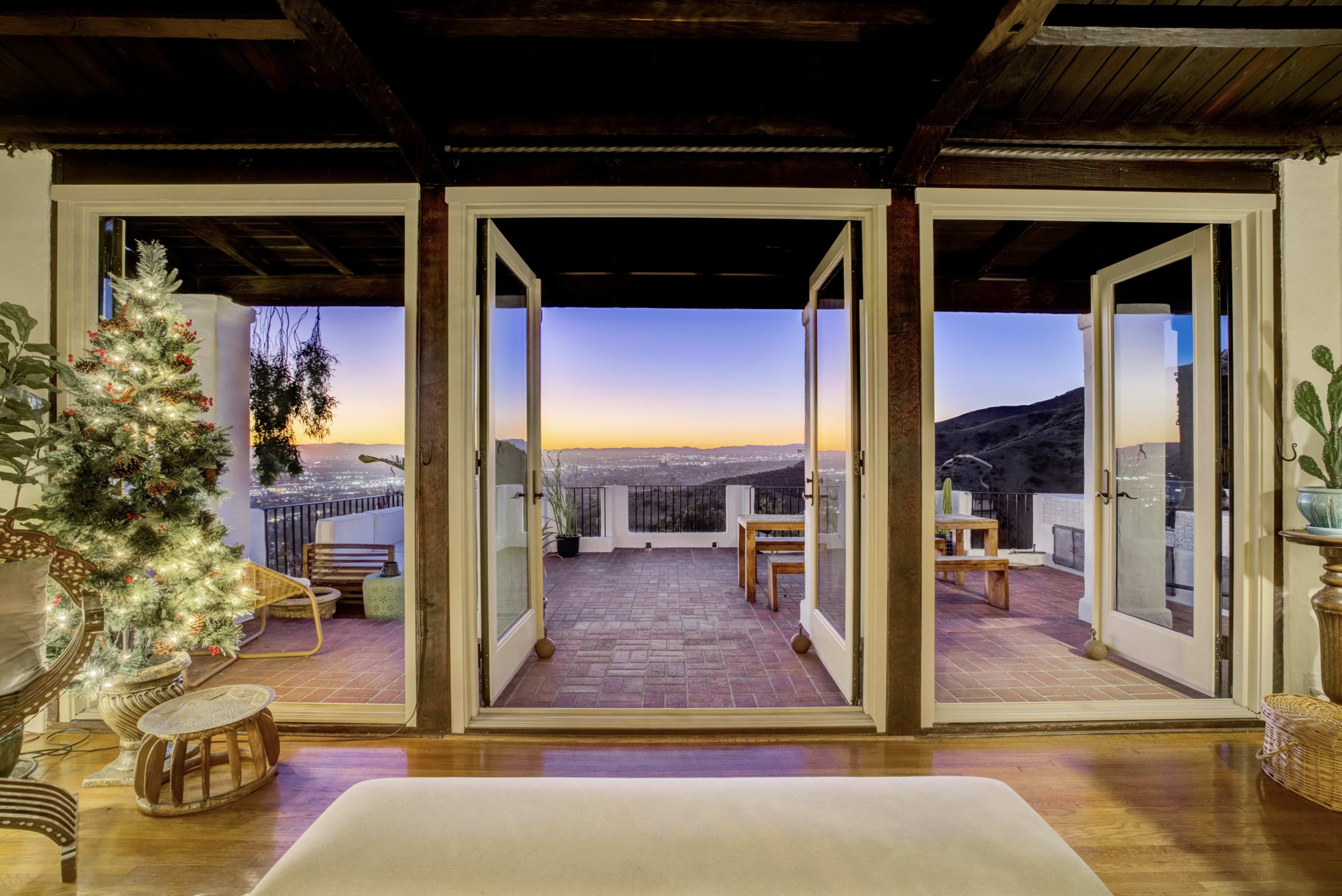 The image shows a cozy interior room with three open glass doors leading to a terrace that features a scenic view of the sunset over distant hills.