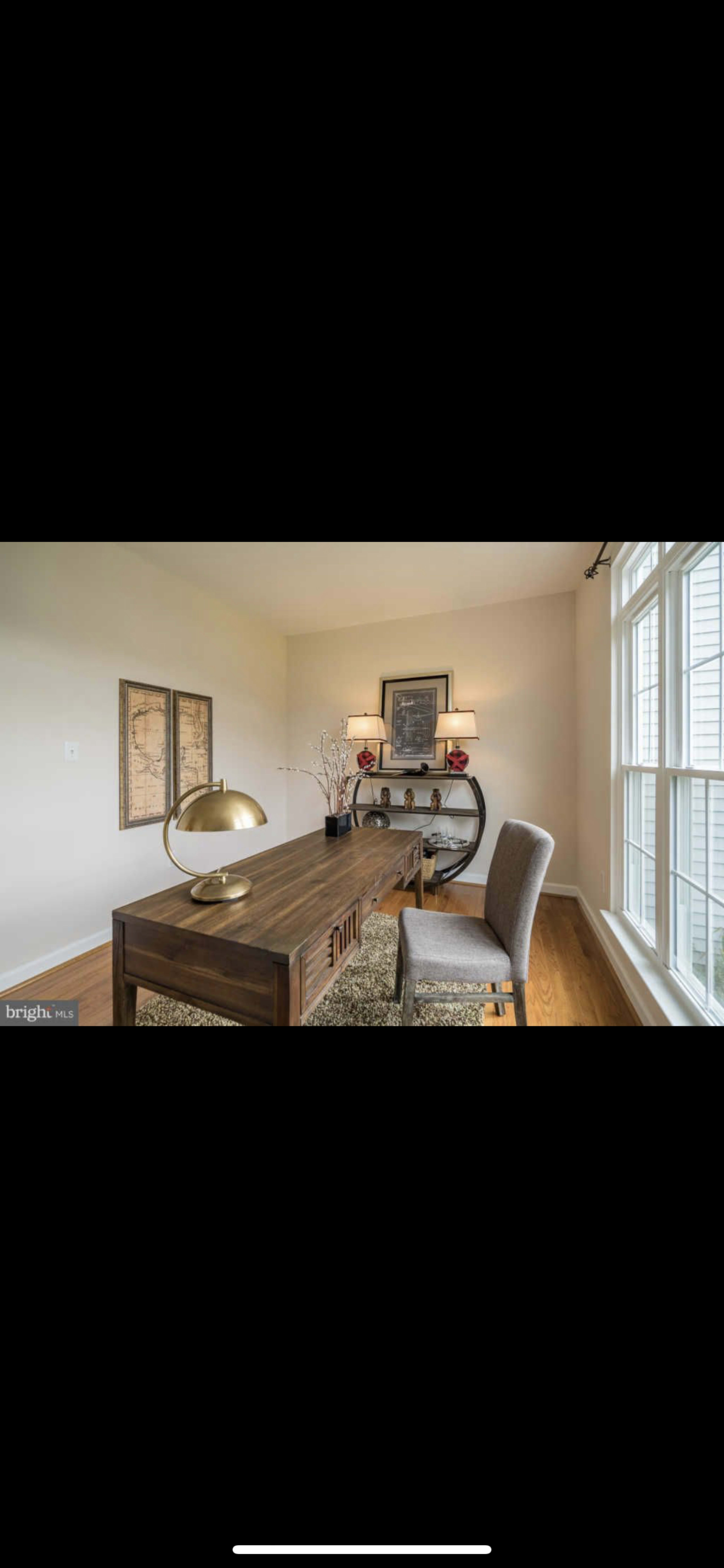 A modern home office features a wooden desk with a lamp, a chair, and decor items against a backdrop of large windows.