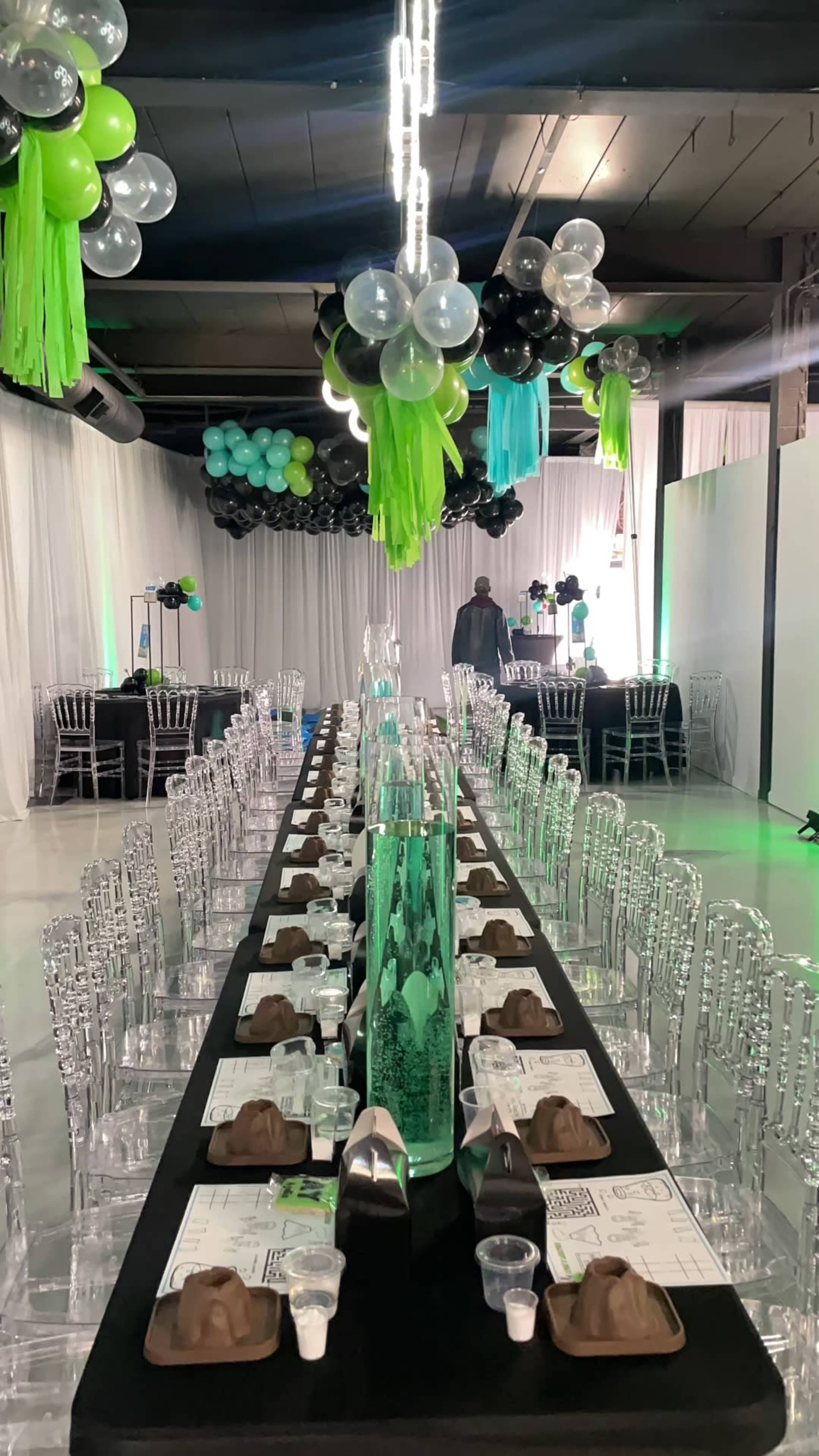 A long banquet table is set with clear chairs and decorative centerpieces, while colorful balloons hang above in an elegantly arranged event space.