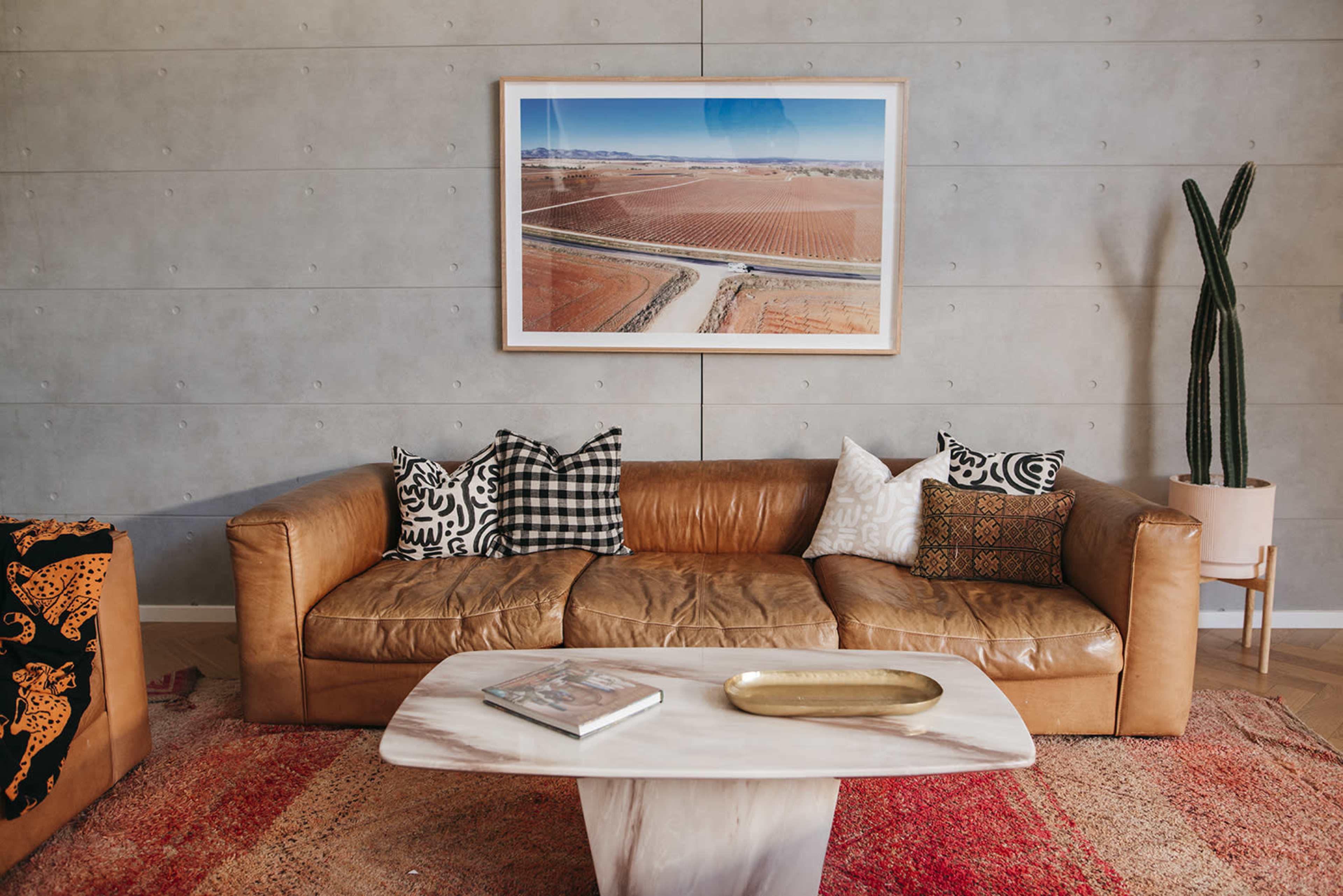 A modern living room featuring a brown leather sofa adorned with patterned pillows, a marble coffee table, and a large framed aerial photograph on the wall above a concrete surface.