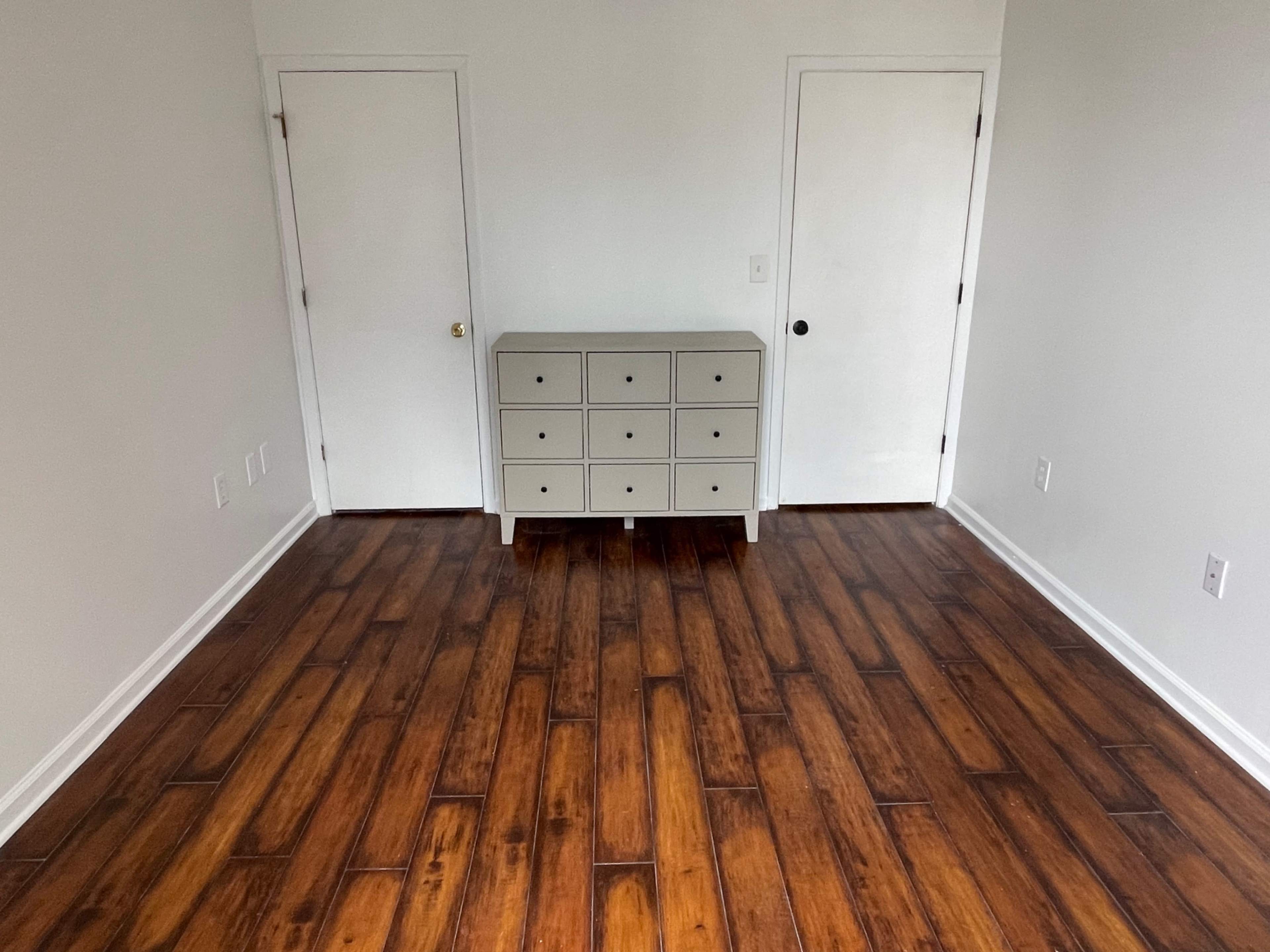 An empty room with wooden flooring, two doors on opposite walls, and a gray dresser positioned in the center.