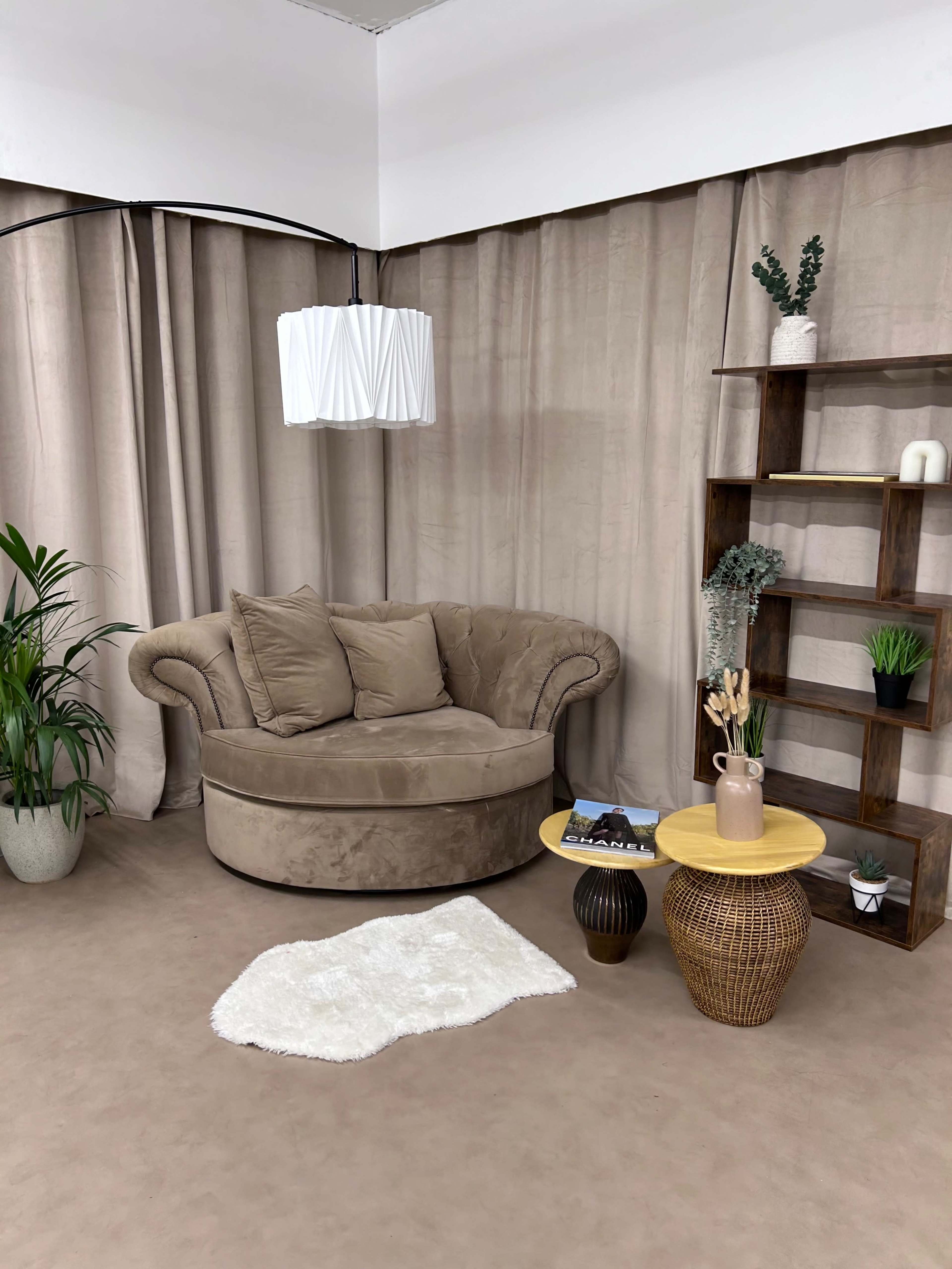 A cozy seating arrangement featuring a circular beige couch, a wooden shelf, and a small round yellow table, all set against a backdrop of neutral-toned curtains.