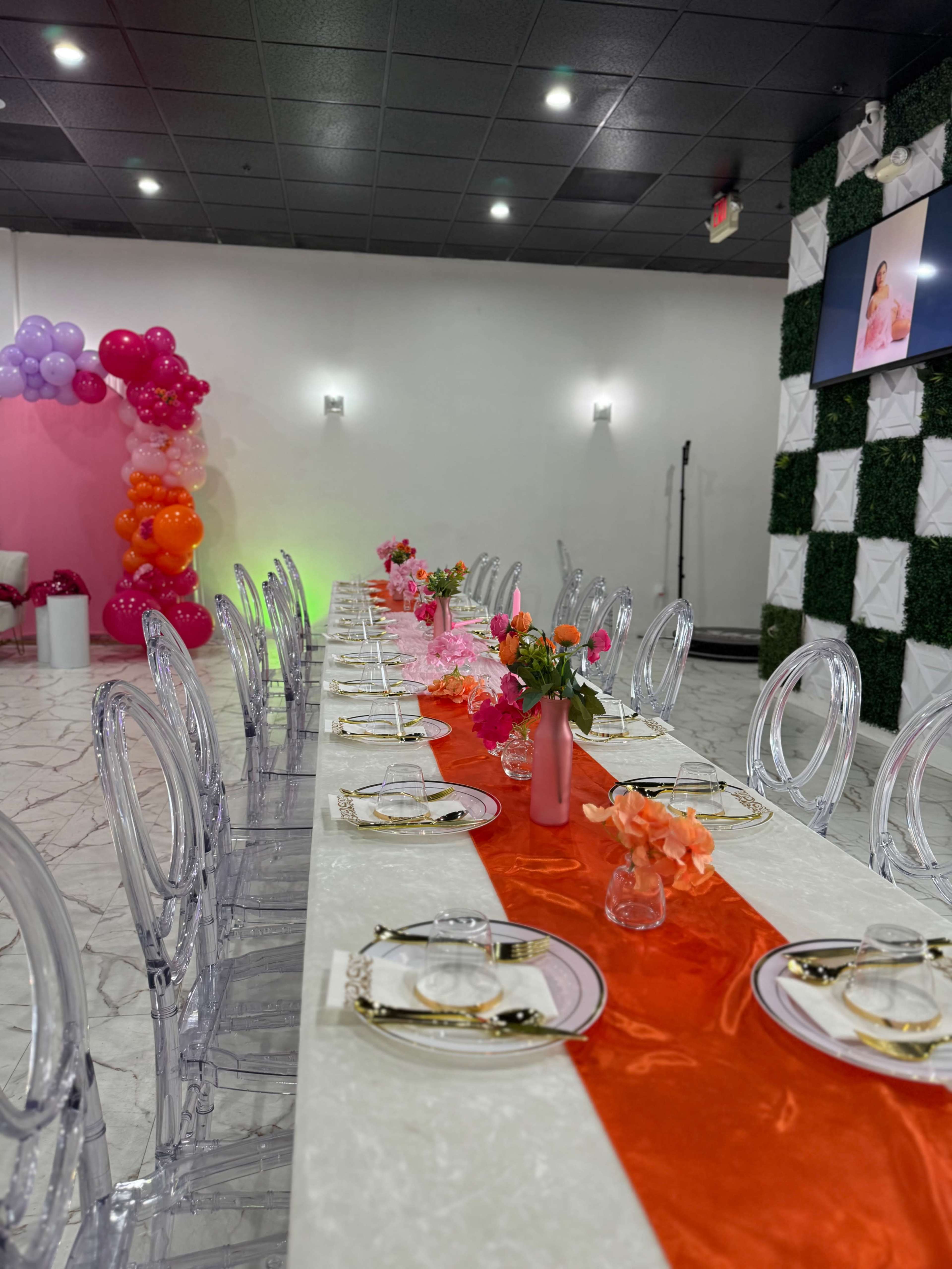 A long dining table is set with clear chairs, a red runner, and colorful floral centerpieces, surrounded by balloons in various hues.