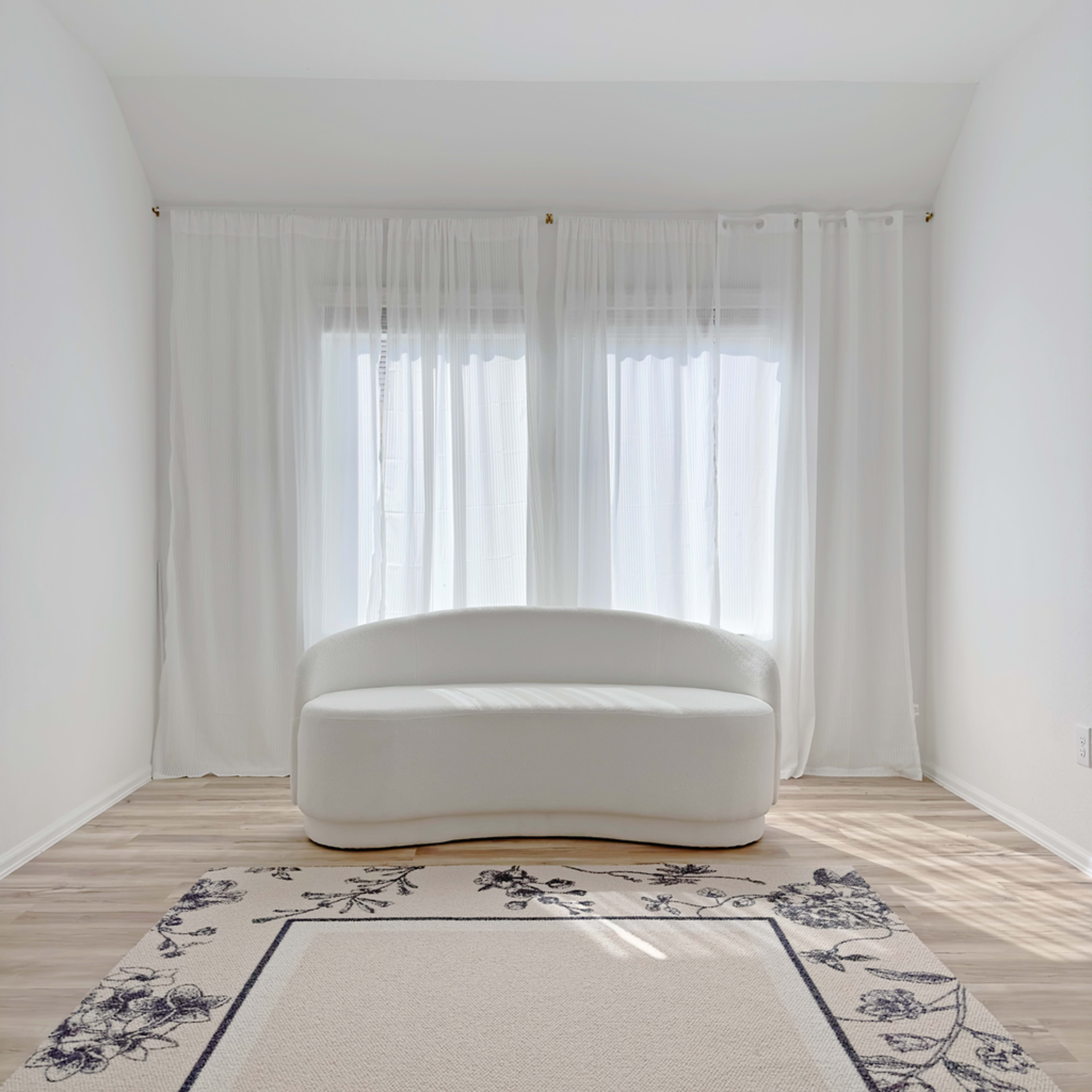 A white curved sofa is positioned in front of large windows draped with sheer white curtains, on a light wood floor with a decorative rug.