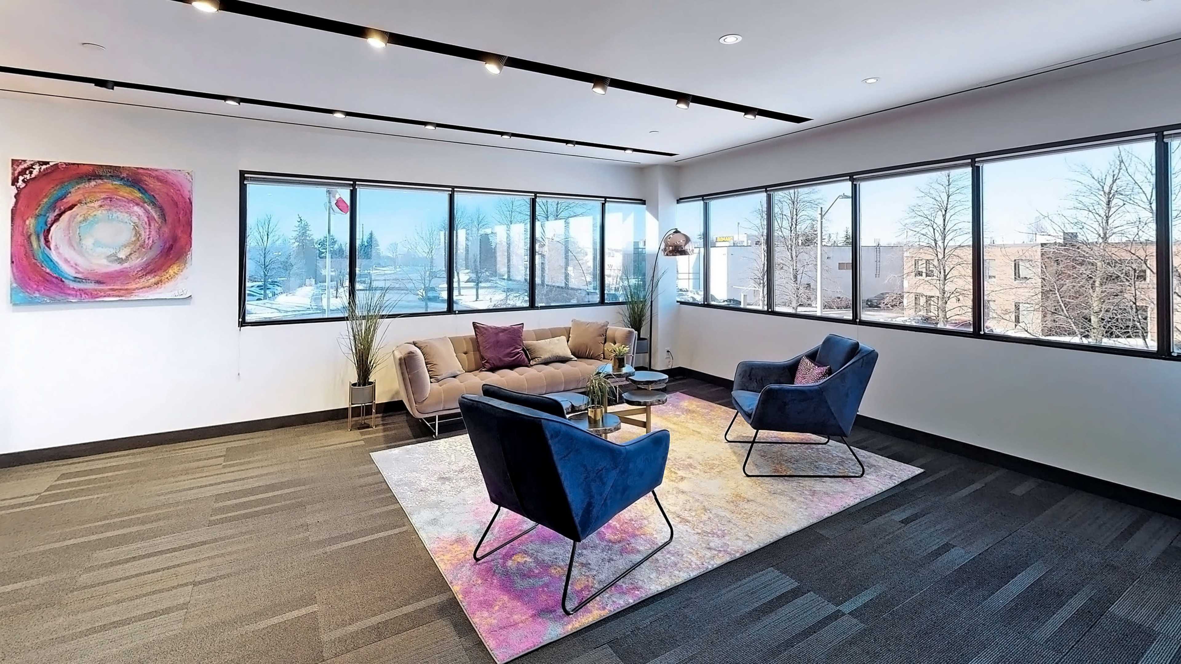 The image shows a modern lounge area with two blue chairs, a beige sofa, a round coffee table, and a colorful rug, all set against large windows that provide a view of an outdoor space.