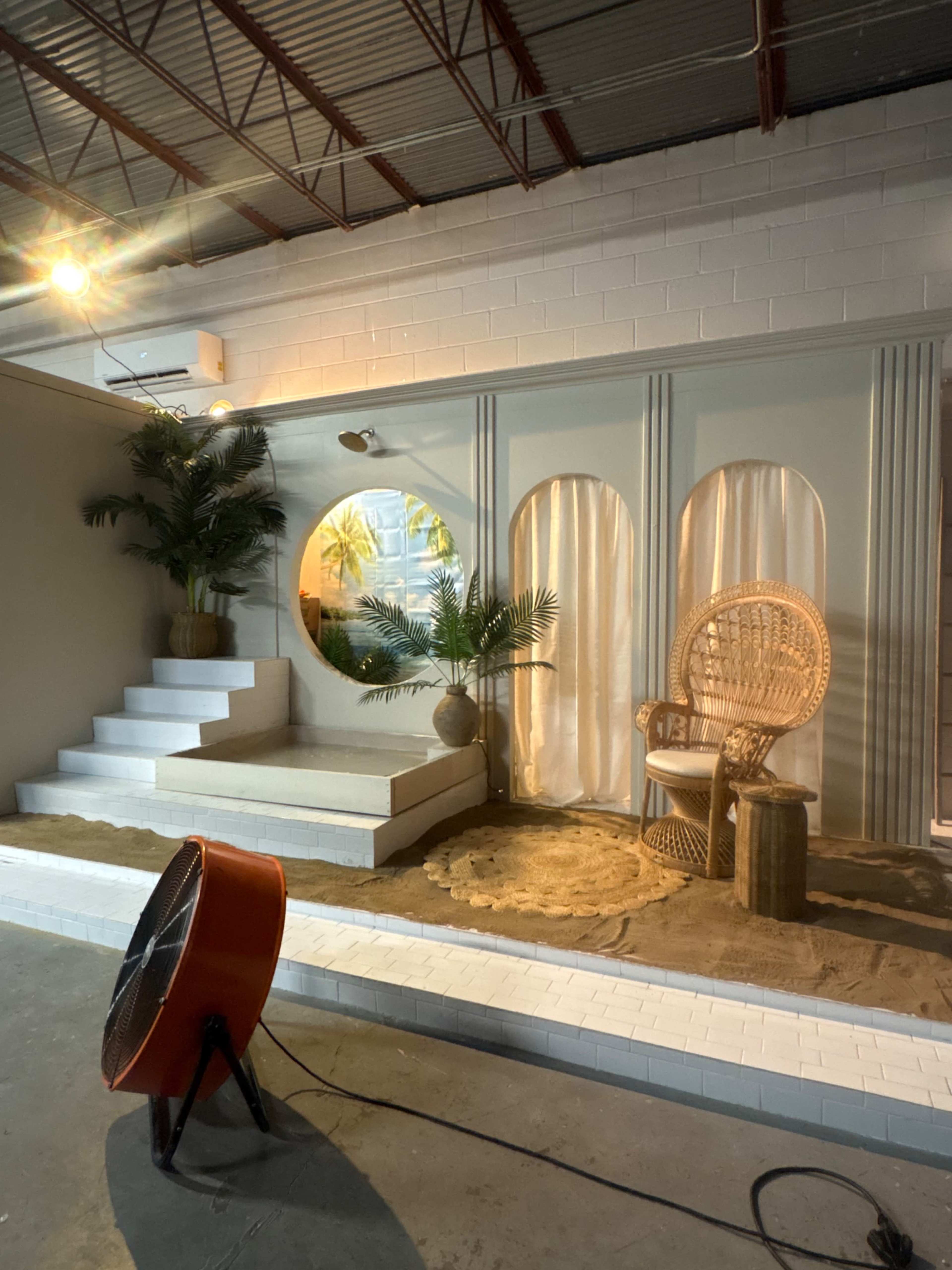 The image shows a minimalist, staged interior featuring steps, a rattan chair, potted plants, and decorative curtains against a light-colored wall.