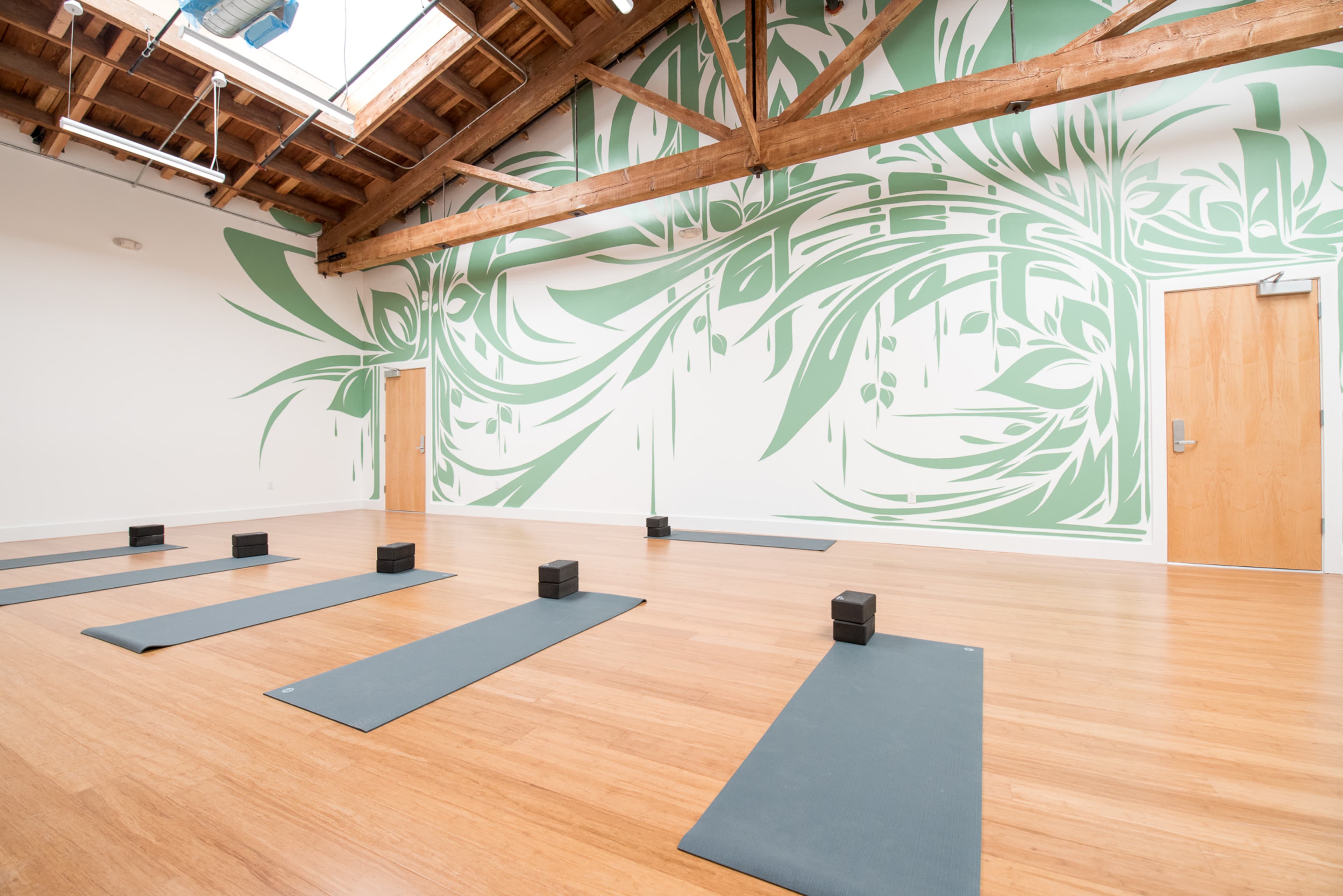 A spacious yoga studio with bamboo flooring, six yoga mats arranged on the floor, and a green mural on one wall.