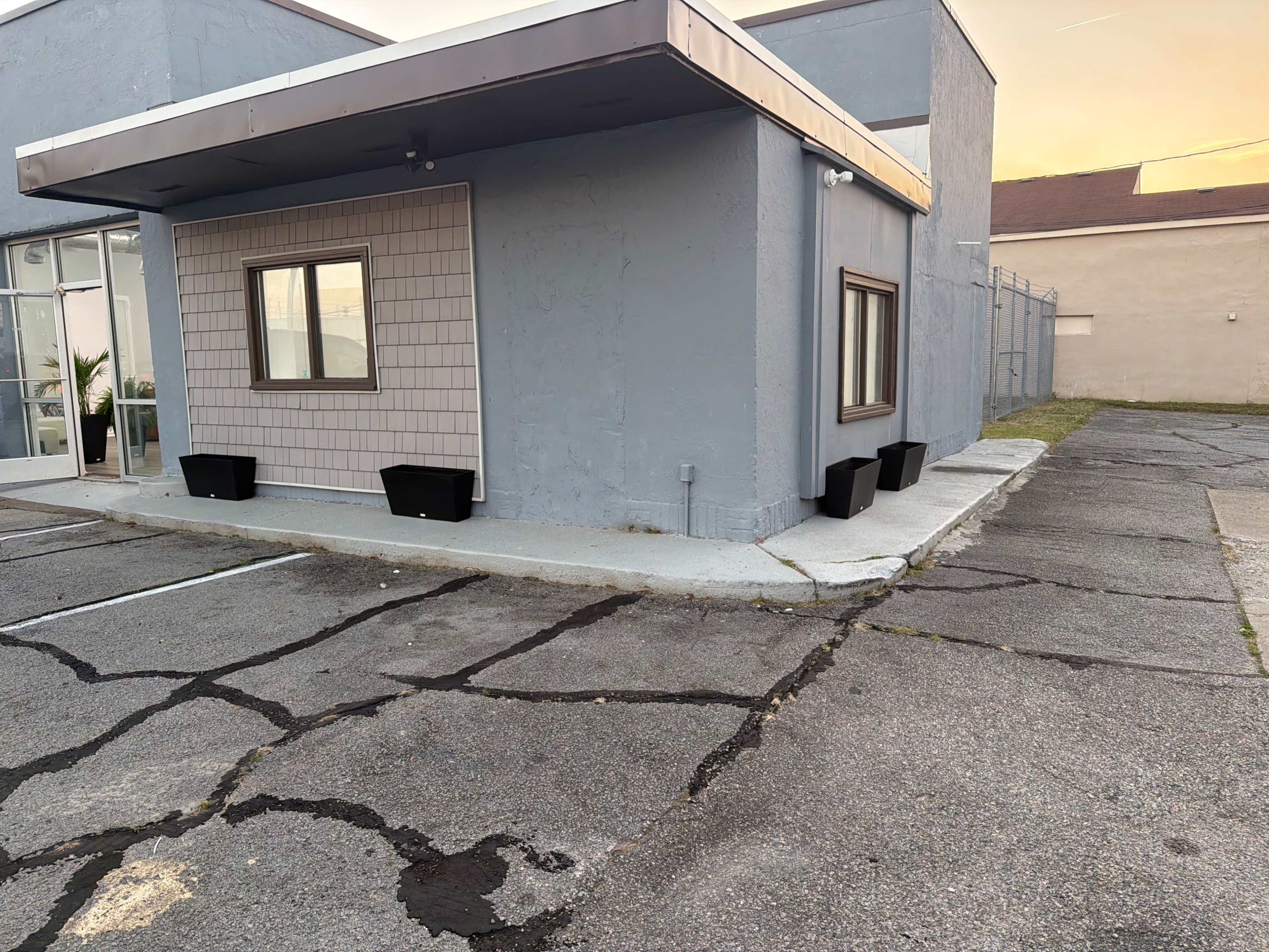 The image shows a small, gray building with a single window and planters, located beside a cracked parking lot.