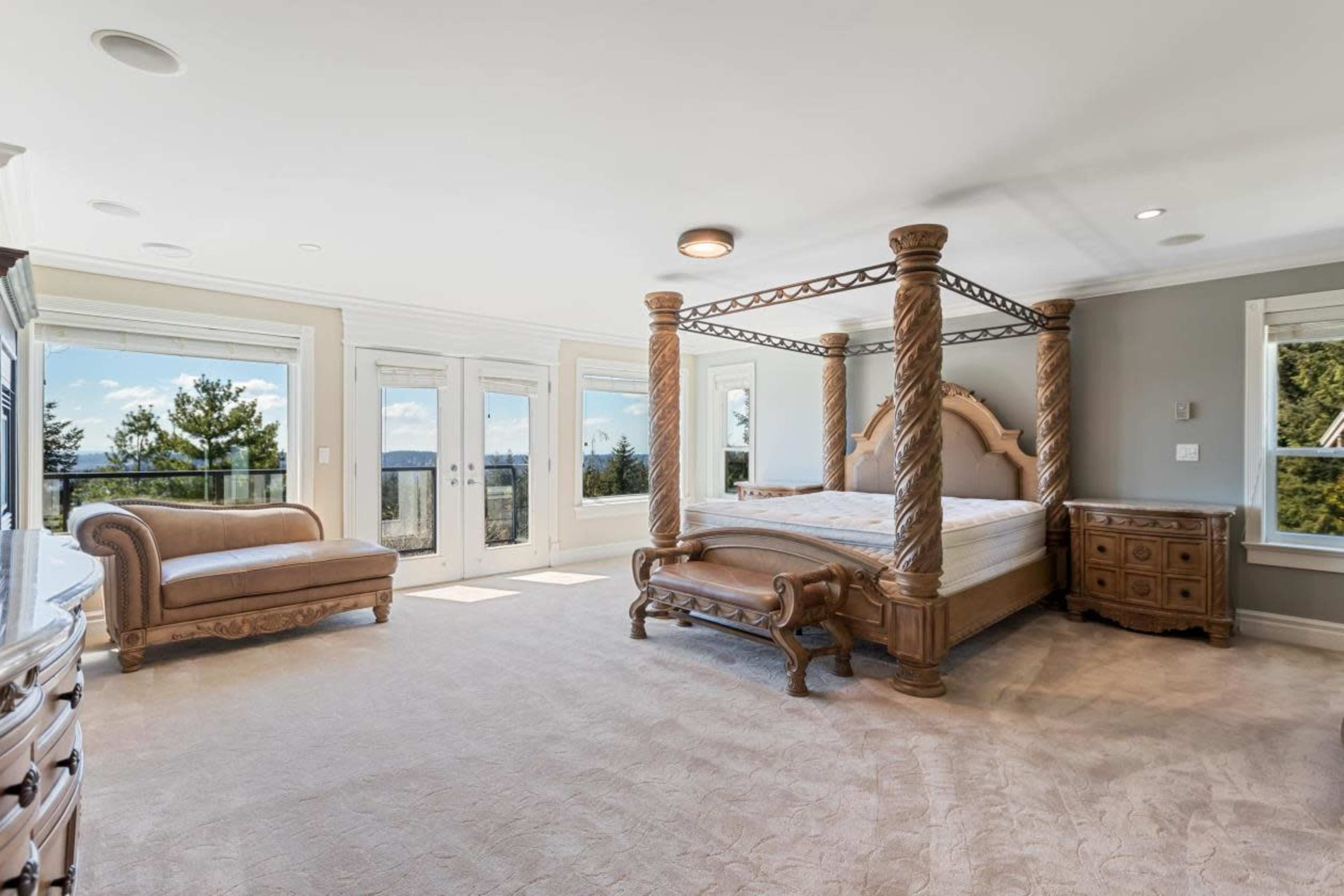 The spacious bedroom features a four-poster bed, a chaise lounge, large windows, and elegant furniture against a backdrop of a scenic view.