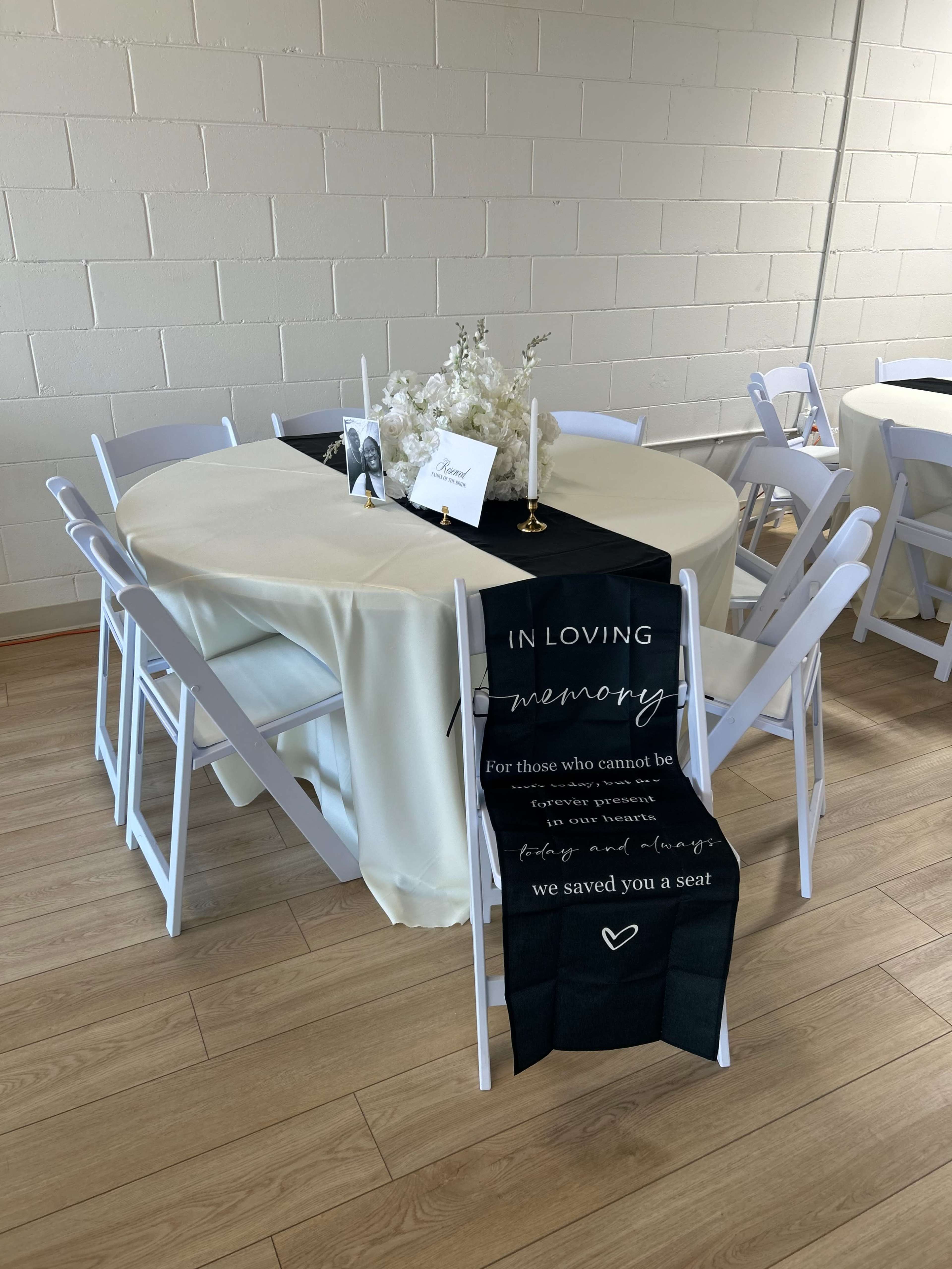 A table is set with a cream-colored tablecloth and a black runner featuring a memorial message, surrounded by white chairs in a bright indoor space.