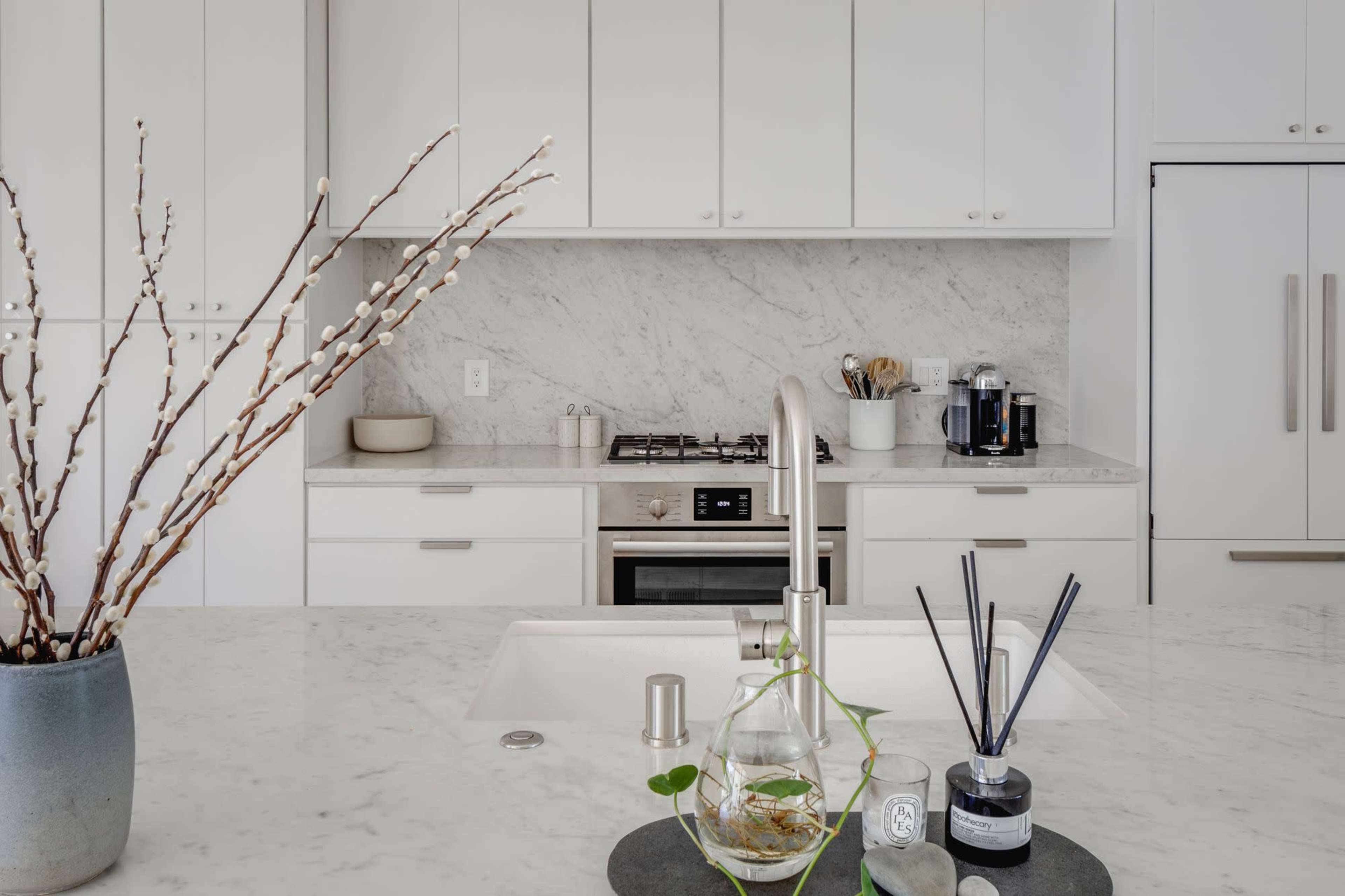 A modern kitchen features white cabinetry, a marble backsplash, a stainless steel gas stove, and a sink with a decorative plant in front.