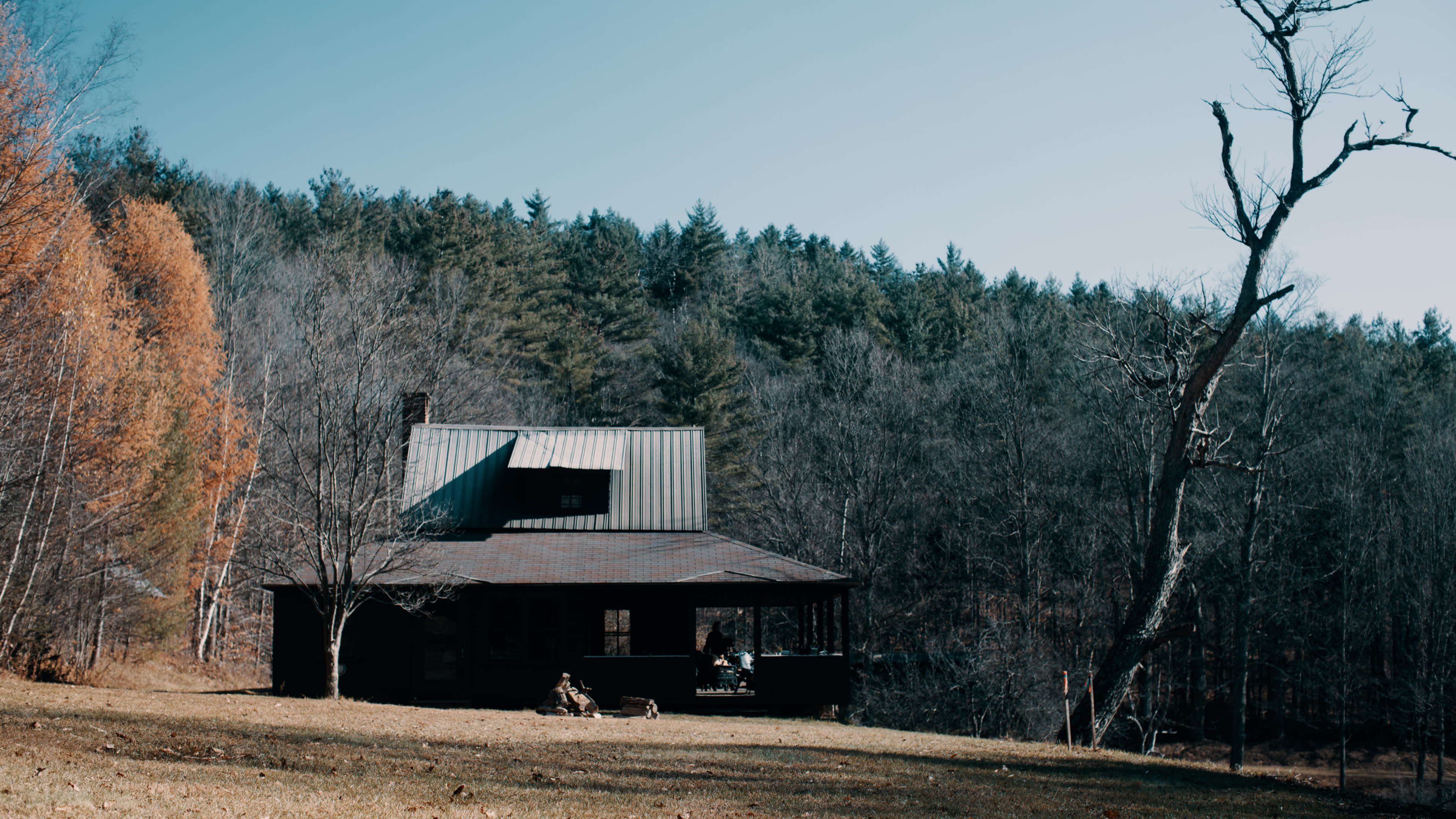 A two-story cabin with a metal roof is surrounded by sparse trees and open grass in a mountainous area.