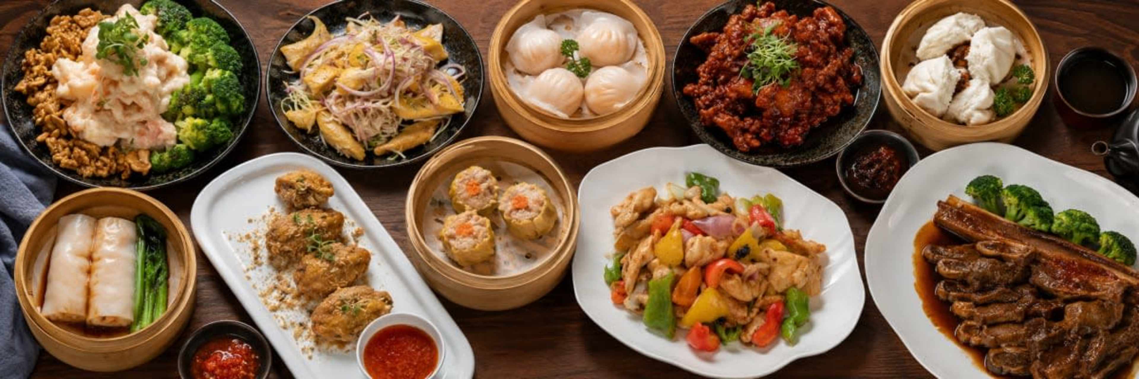 The image displays a variety of Asian dishes arranged on a wooden table, including dumplings, stir-fried chicken, and vegetable platters.