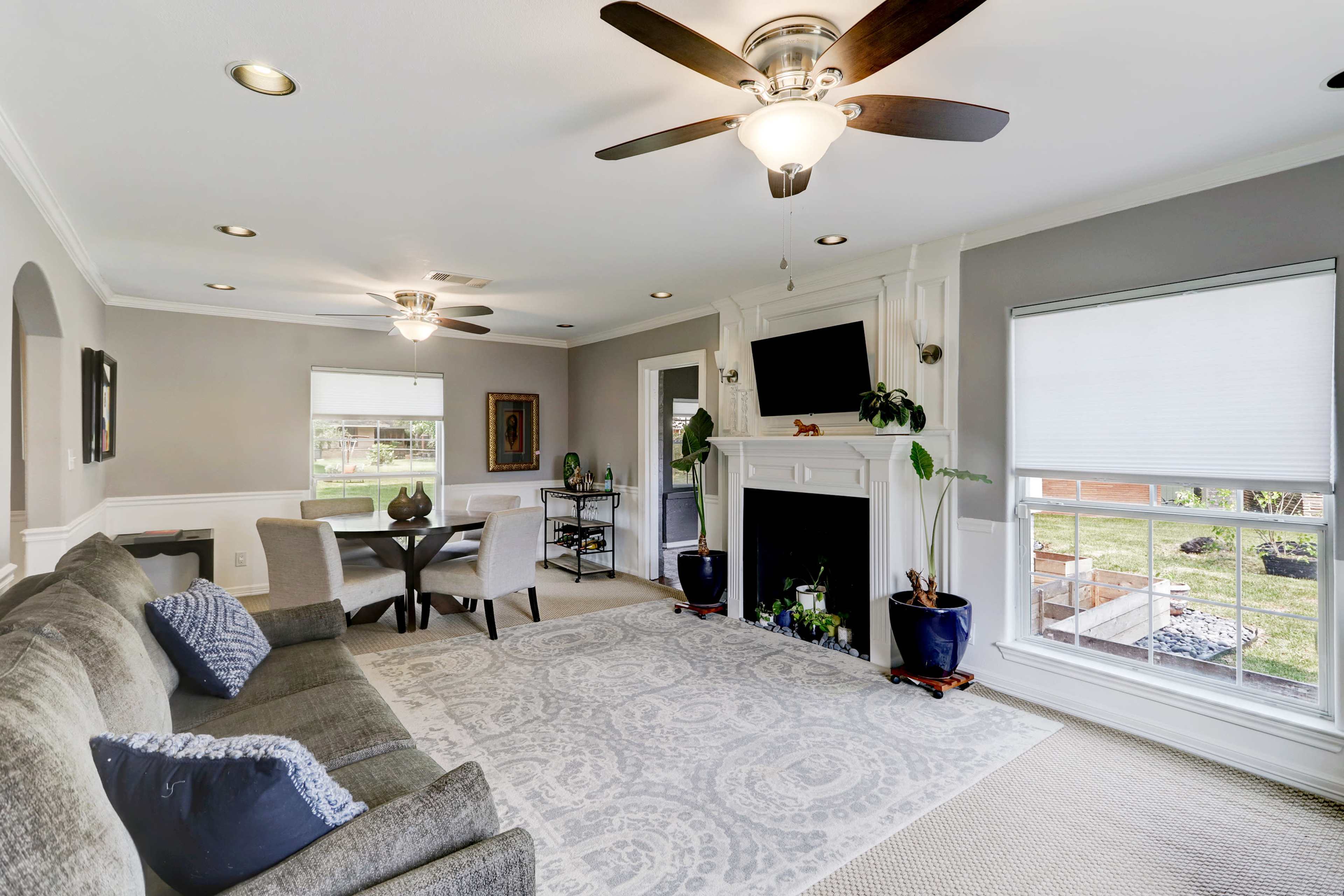 A living room with a gray and white color scheme, featuring a seating area, a fireplace, and a view of a backyard through large windows.