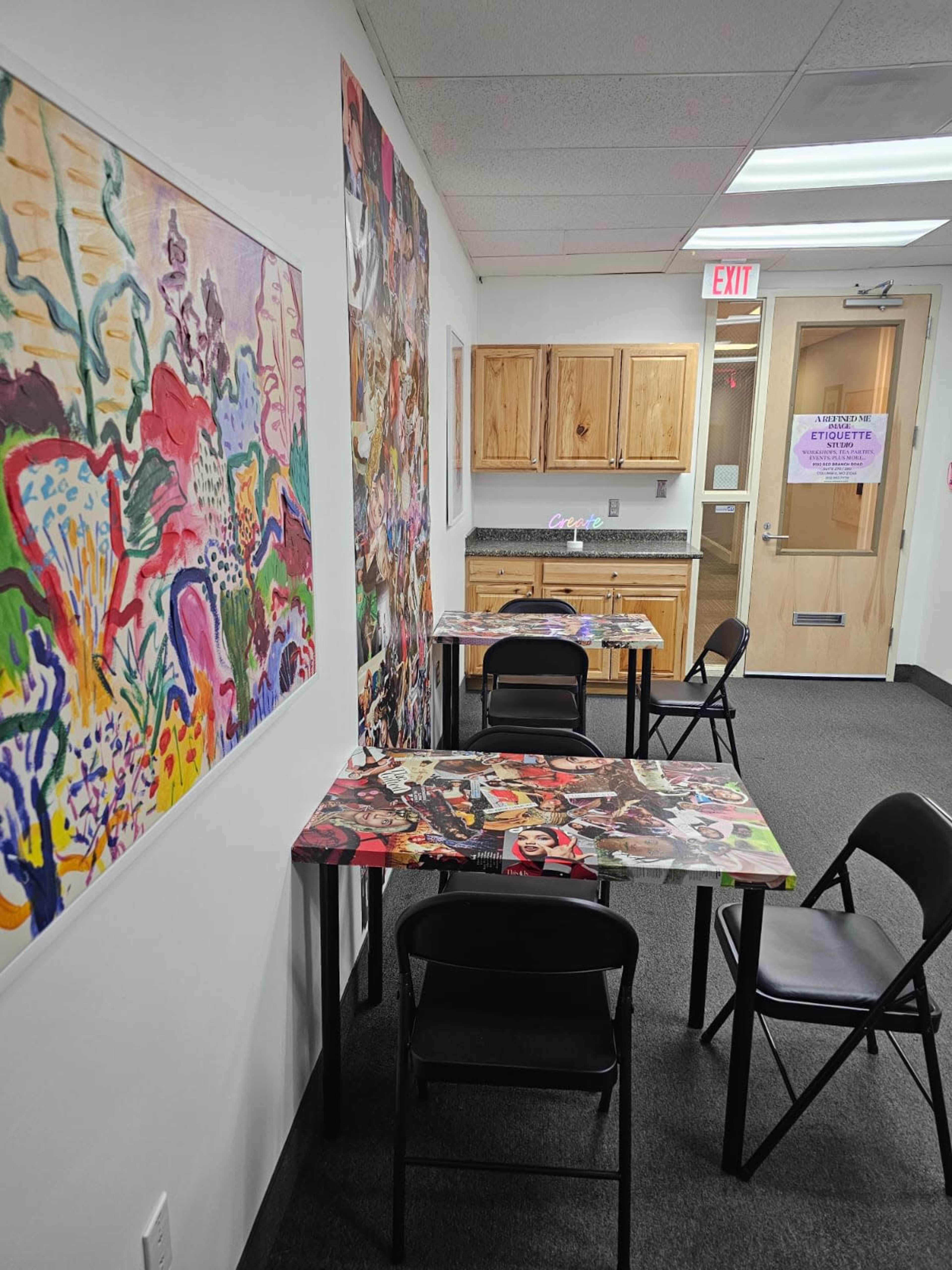 A narrow hallway with four tables covered in artwork and chairs, alongside colorful wall art and a kitchenette area in the background.