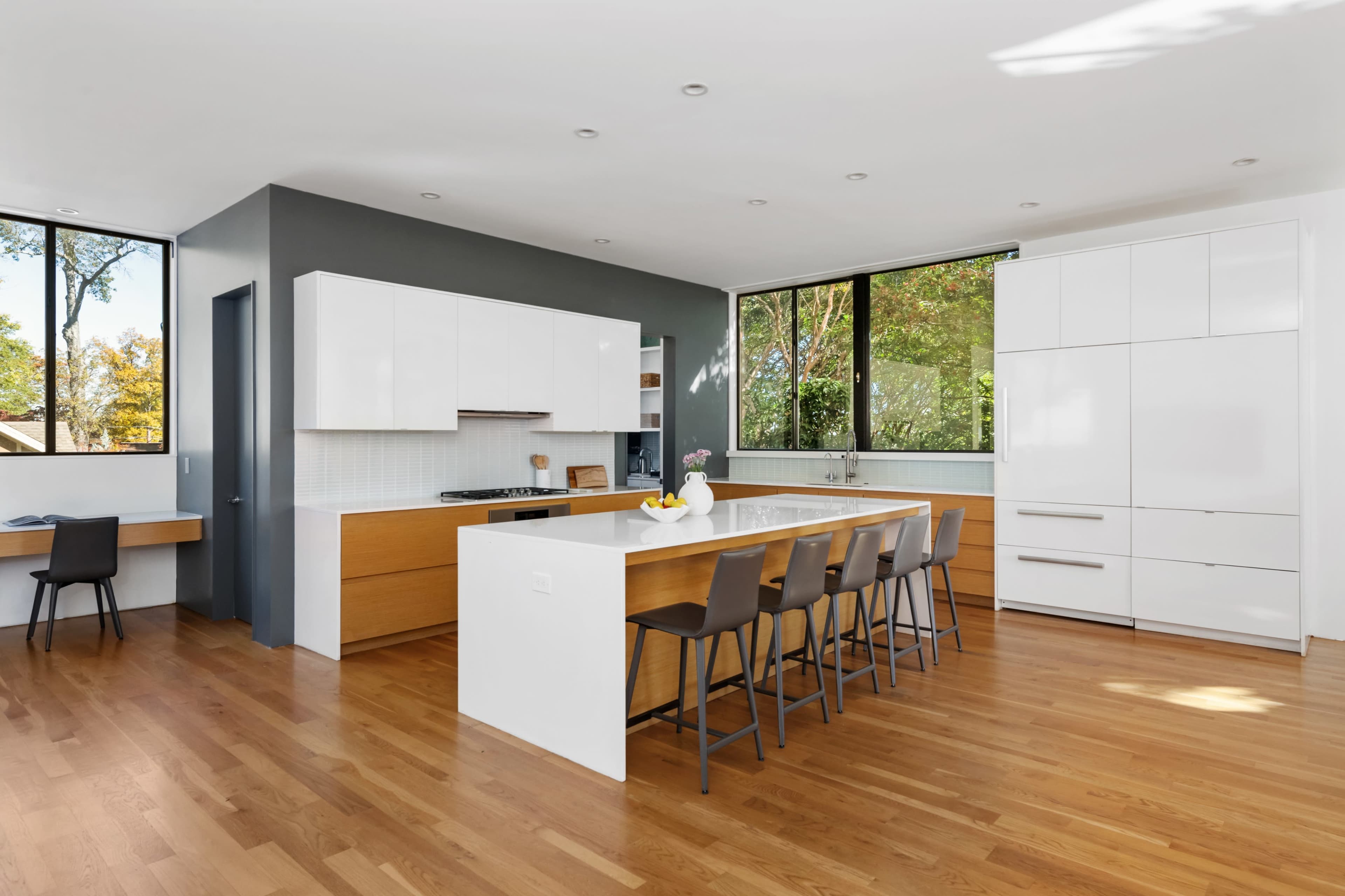 The image features a modern kitchen with a central island and bar seating, large windows providing natural light, and a minimalist design in neutral colors.