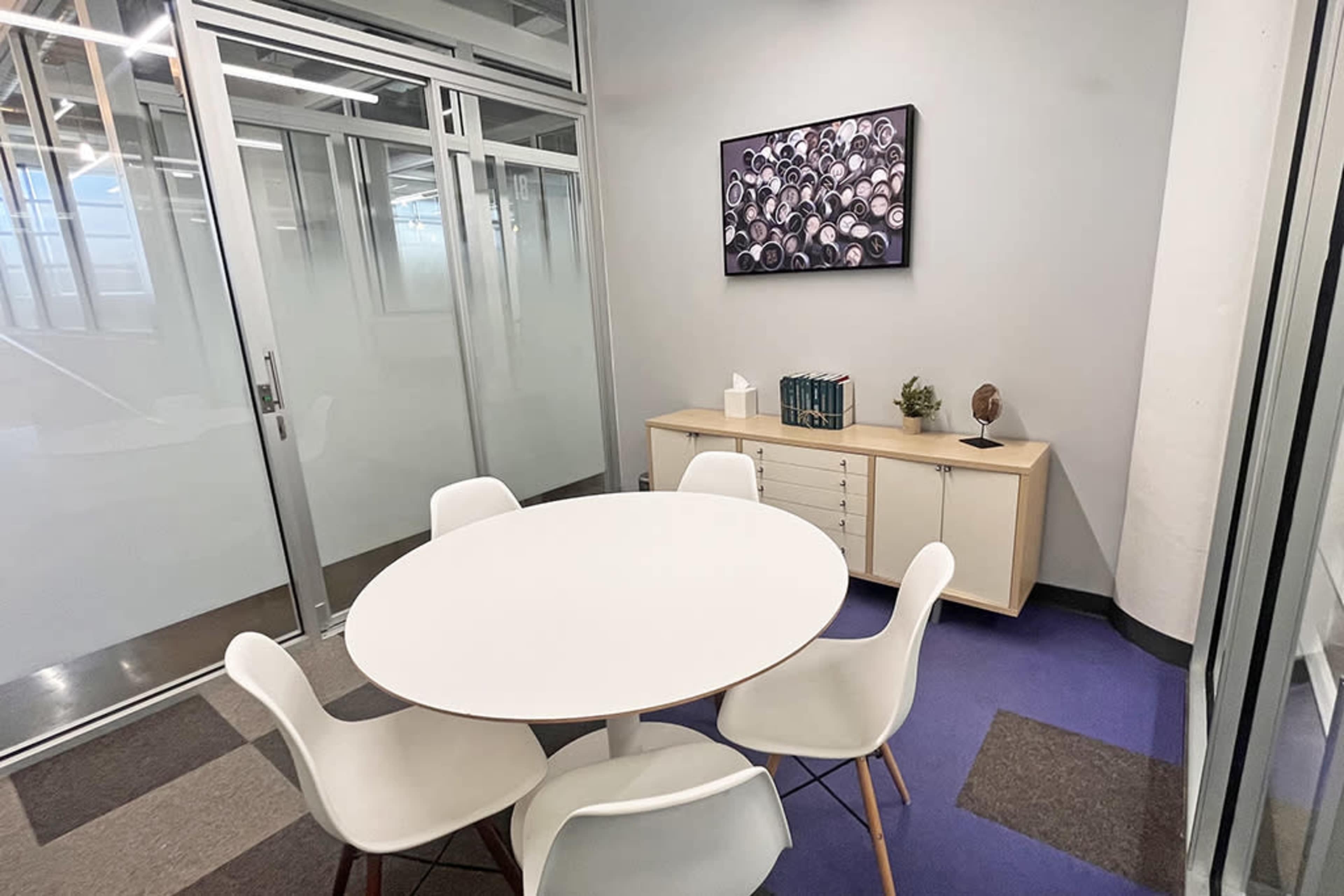 The image shows a small conference room with a round white table and four white chairs, featuring a cabinet against the wall and a framed artwork above it.