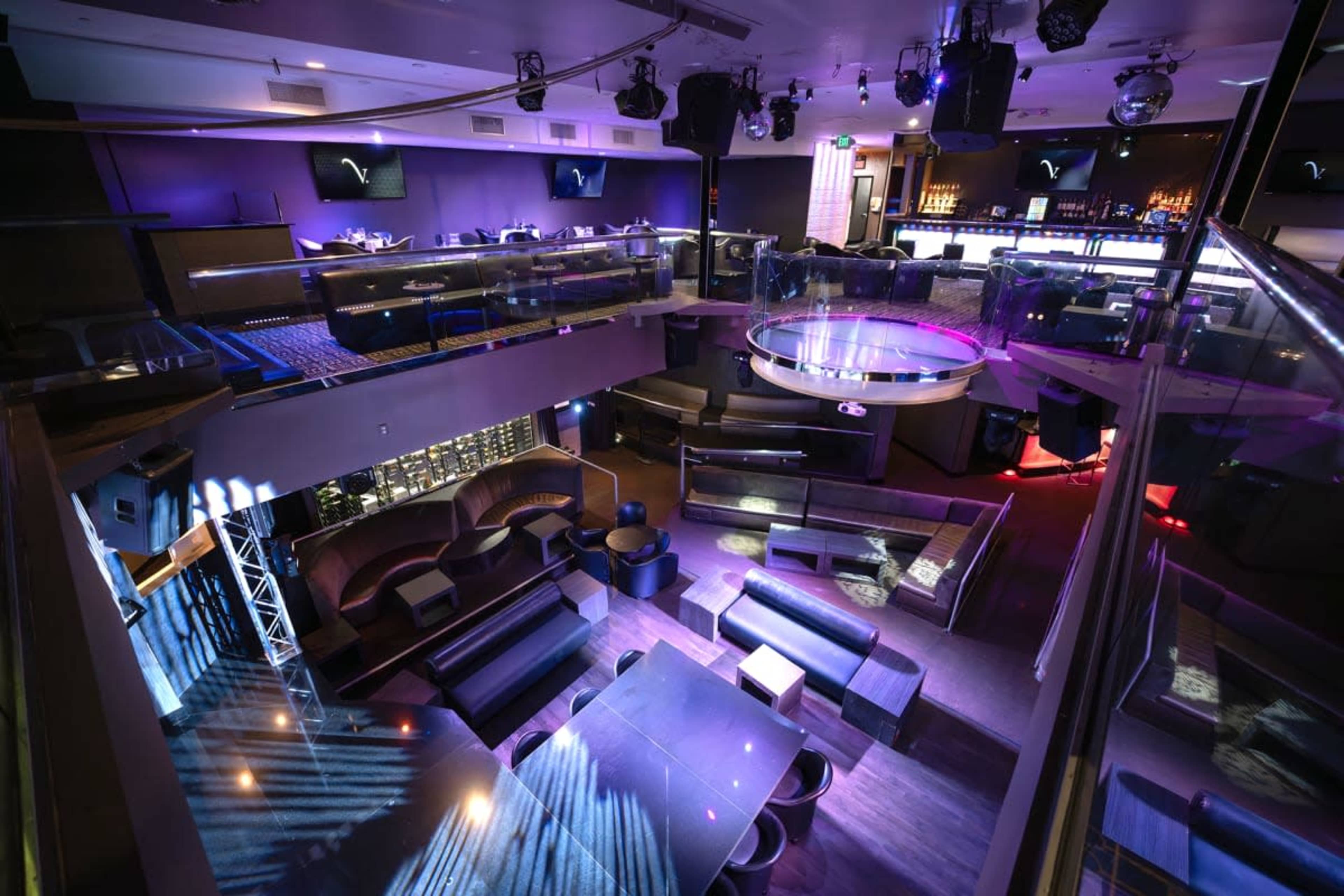 The image depicts an upscale nightclub interior with a central circular stage, tiered seating areas, and a bar counter illuminated by colorful lights.