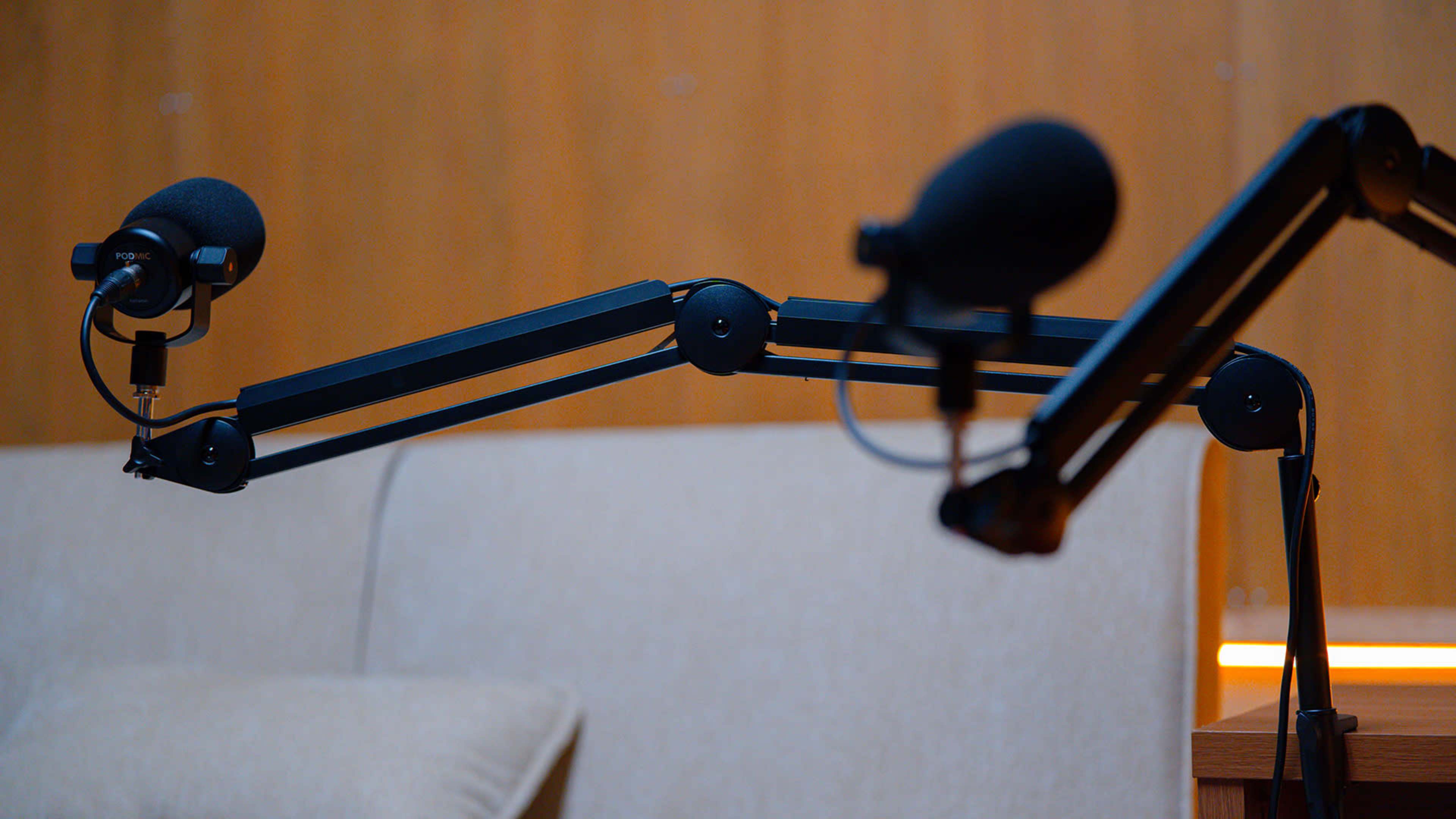 The image shows two microphone arms with attached microphones positioned near a beige couch against a wooden background.